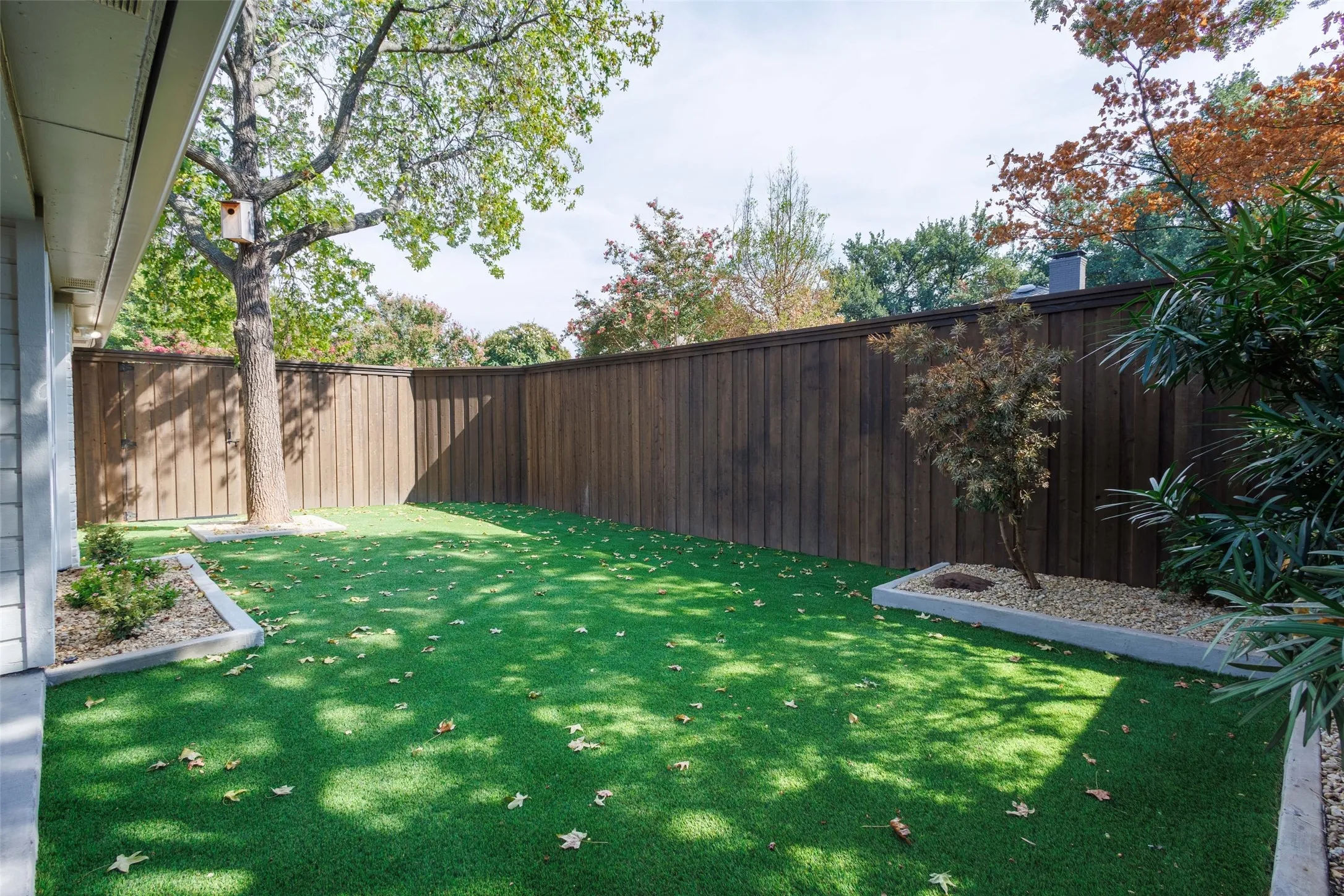 Expansive backyard featuring artificial turf, tall wood fencing, established trees, and raised landscape beds with gravel