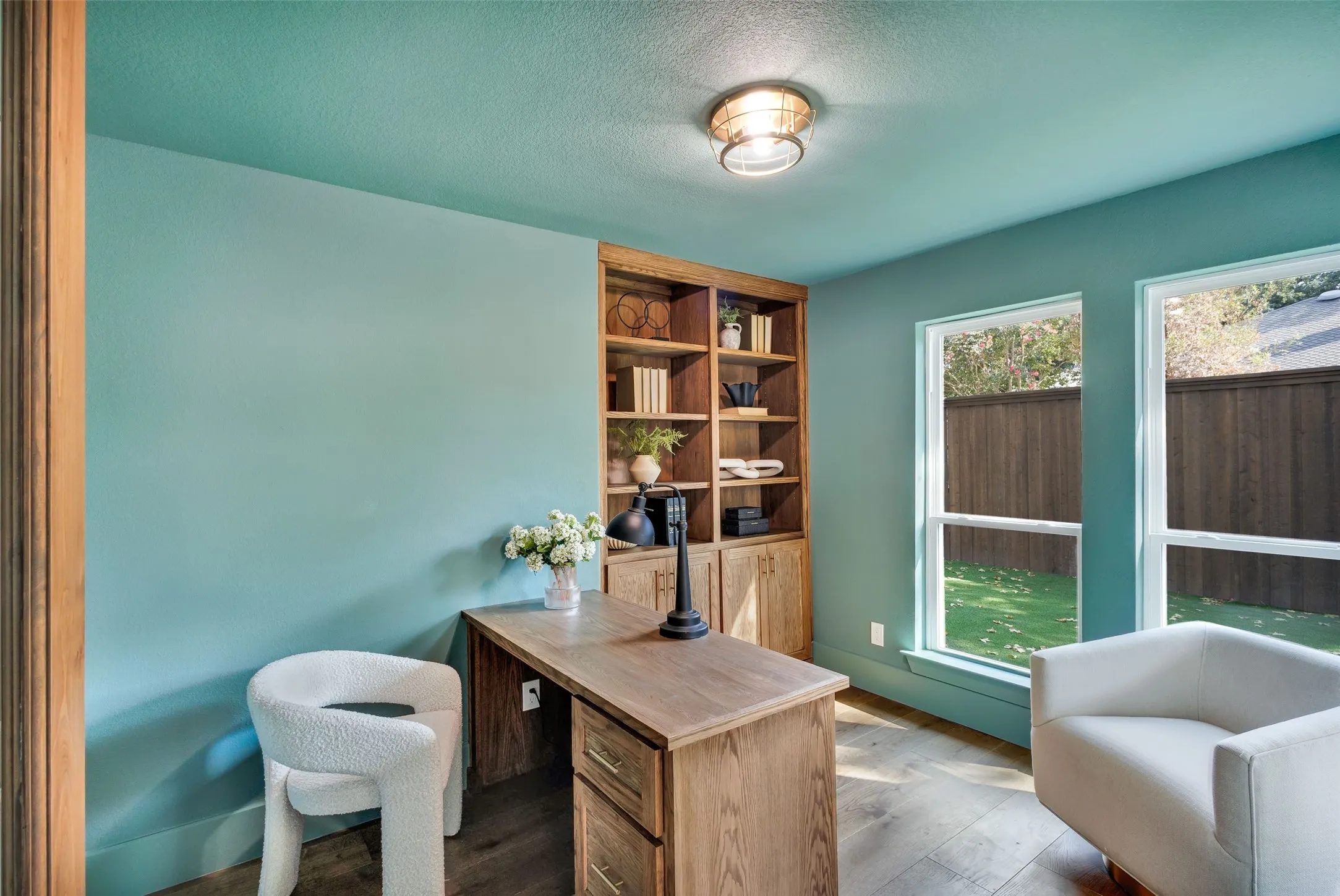 Wood-finish flooring, built-in shelving, two large windows, wood desk with drawers, and a ceiling-mounted light fixture