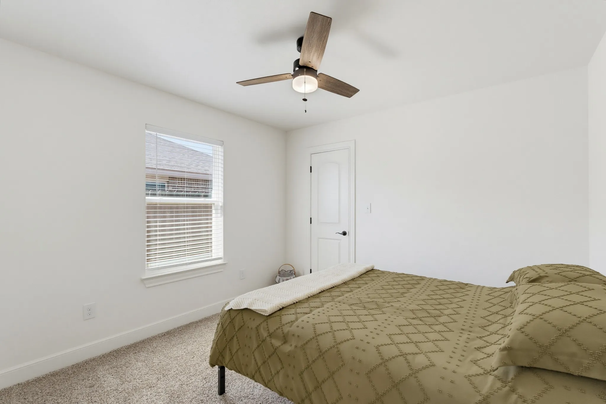 Neutral white walls, window with blinds, wood-finish ceiling fan with integrated lighting, door with dark hardware, and carpeted flooring