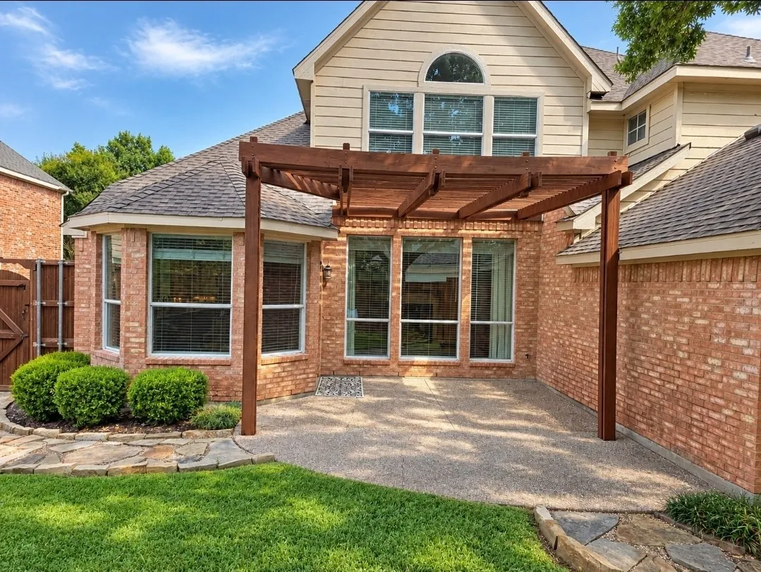 Brick exterior with a wood pergola, expansive windows, exposed aggregate patio, and a stone-lined garden bed