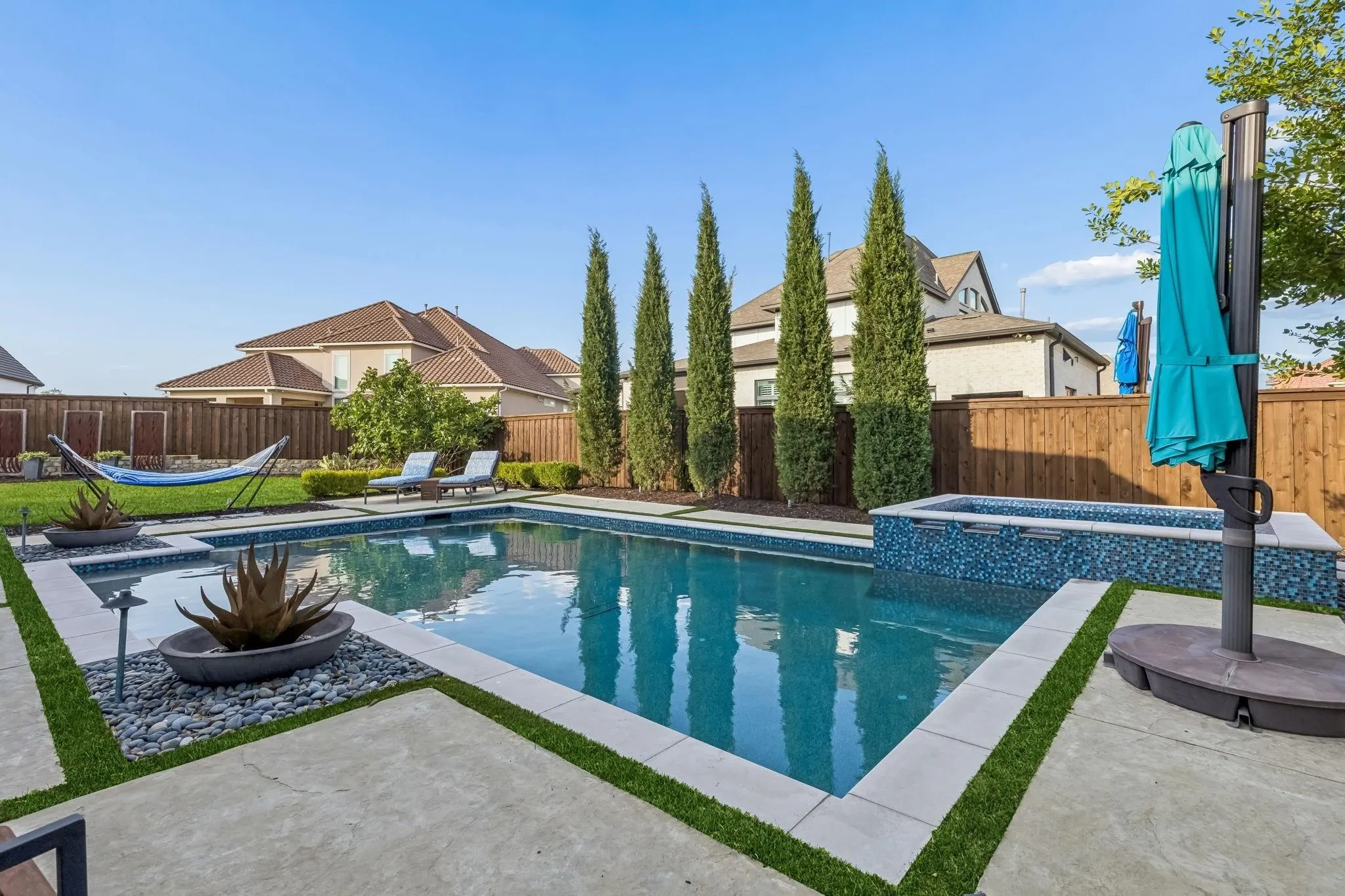 Backyard featuring a rectangular swimming pool with integrated spa, concrete hardscaping with artificial turf accents, and mature trees providing privacy
