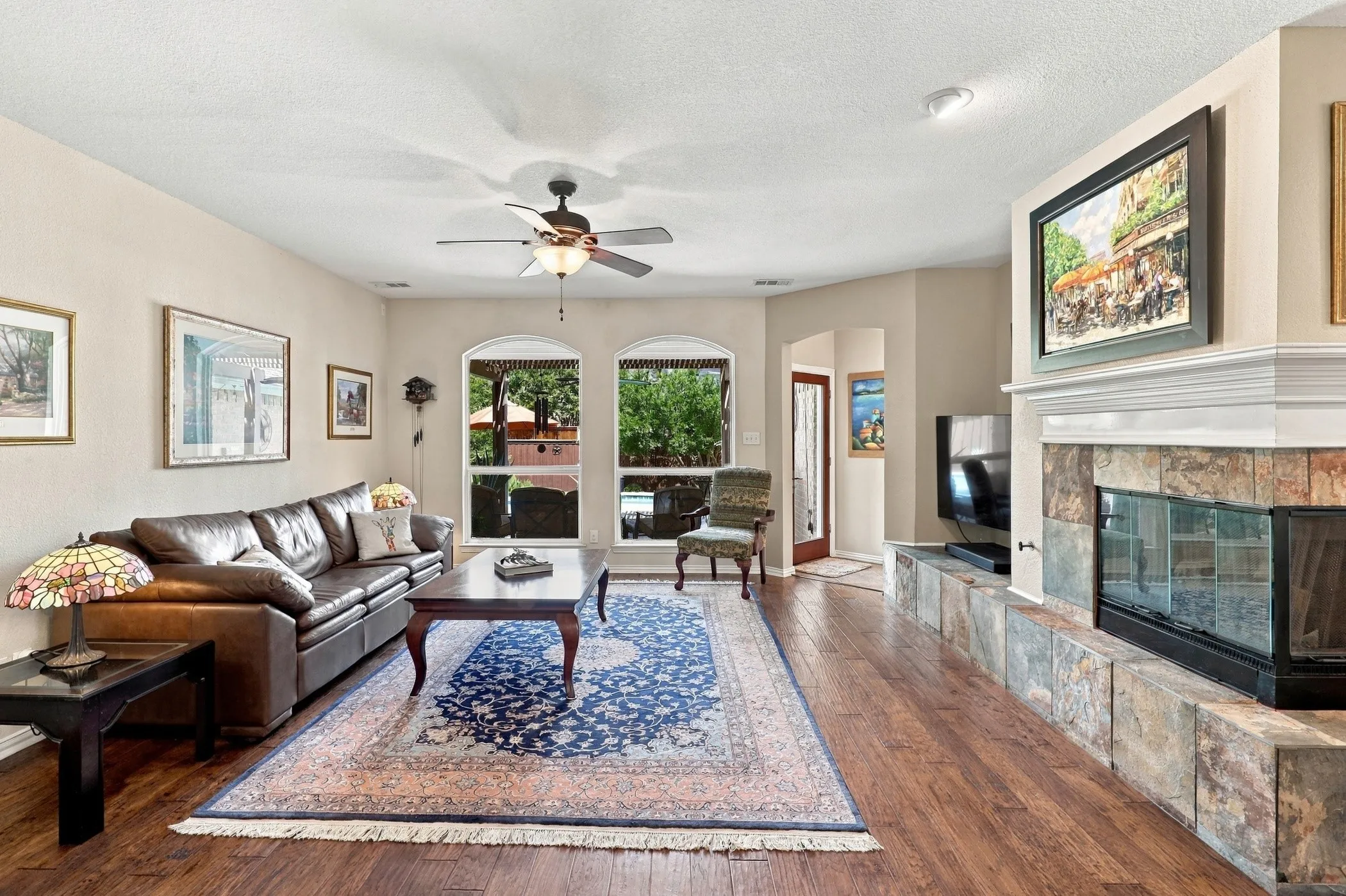 Overlooking backyard pool.  Wood-finish flooring, decorative slate corner fireplace with white mantel, arched windows, ceiling fan with integrated lighting, and neutral wall tones.