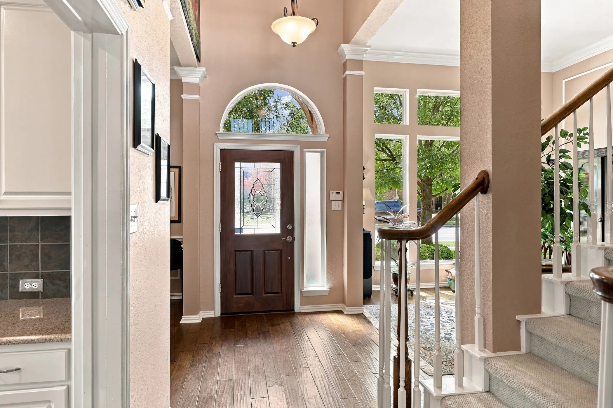 Wood-finish flooring, a front entry door with leaded glass, an arched transom window, a staircase with carpeted treads and white balusters, and a built-in kitchen counter with backsplash tile