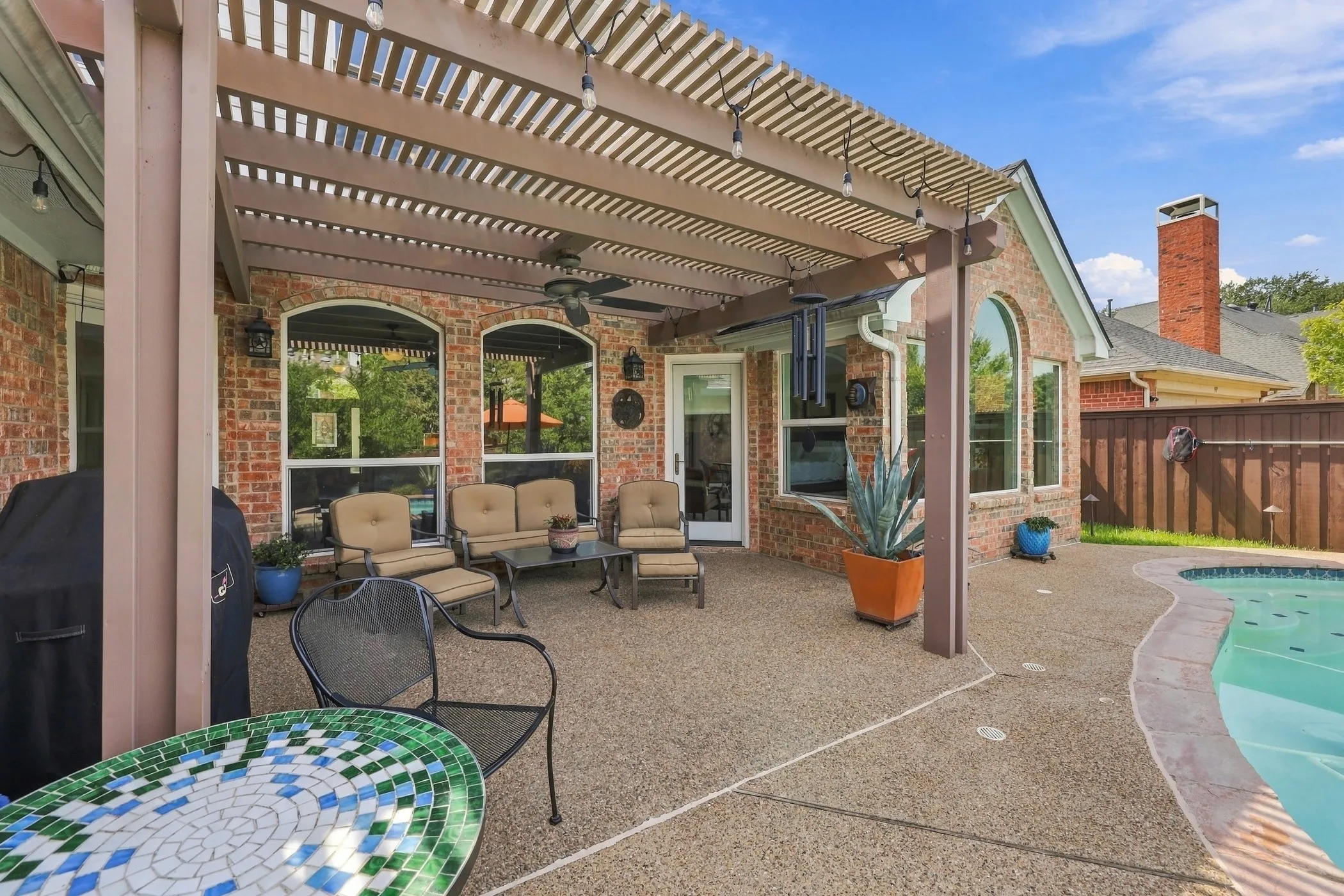 Expansive outdoor living area featuring a covered patio with a slatted pergola, exposed aggregate concrete patio, and curved swimming pool.  Recently re-stained fence.