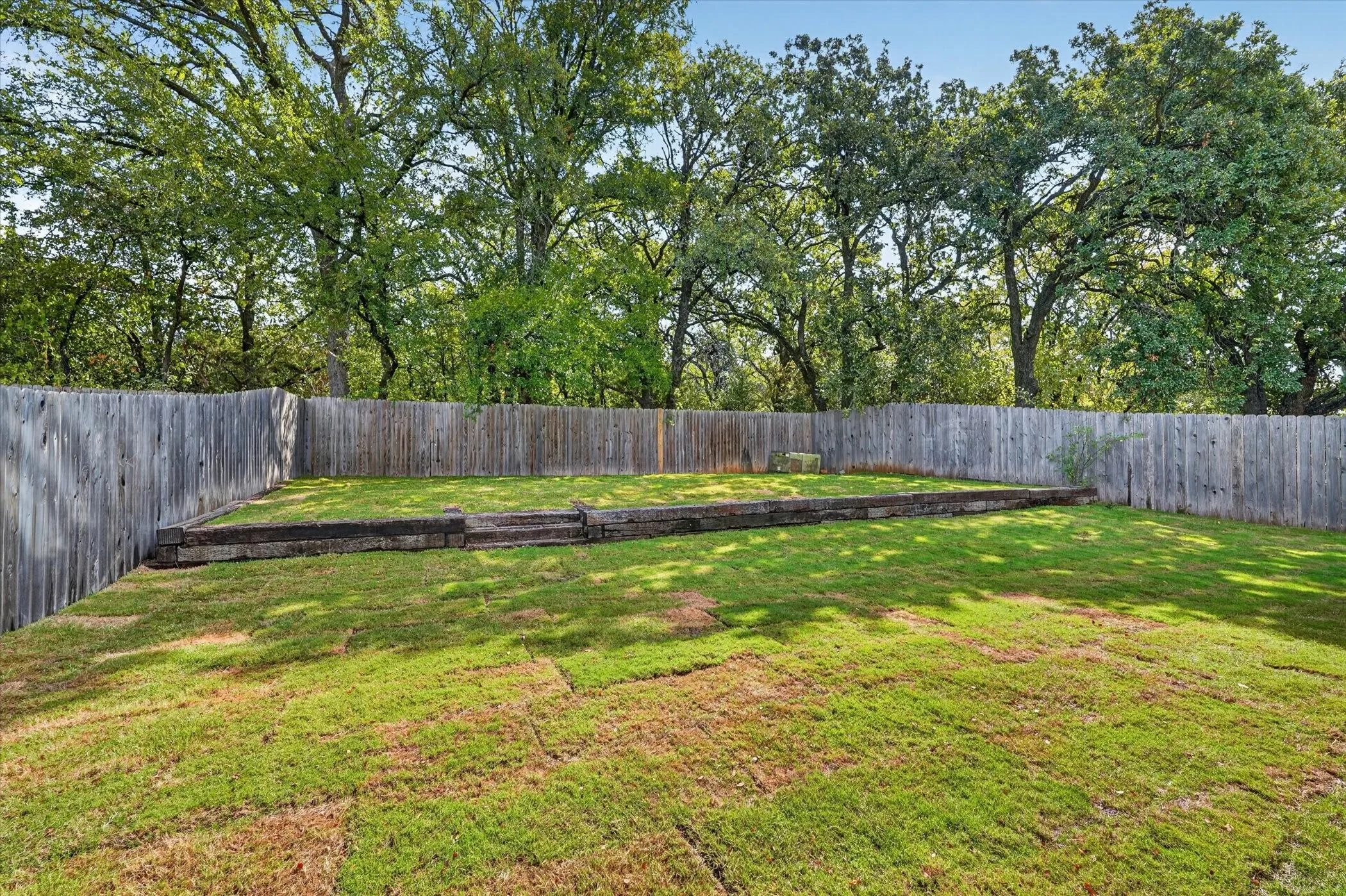 Fenced backyard featuring a tiered lawn, wood retaining wall, and mature perimeter trees