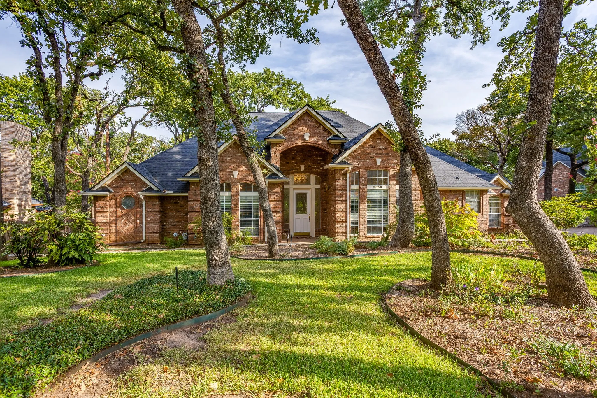 Brick exterior, dark shingle roof, mature trees, arched entryway, and bay window