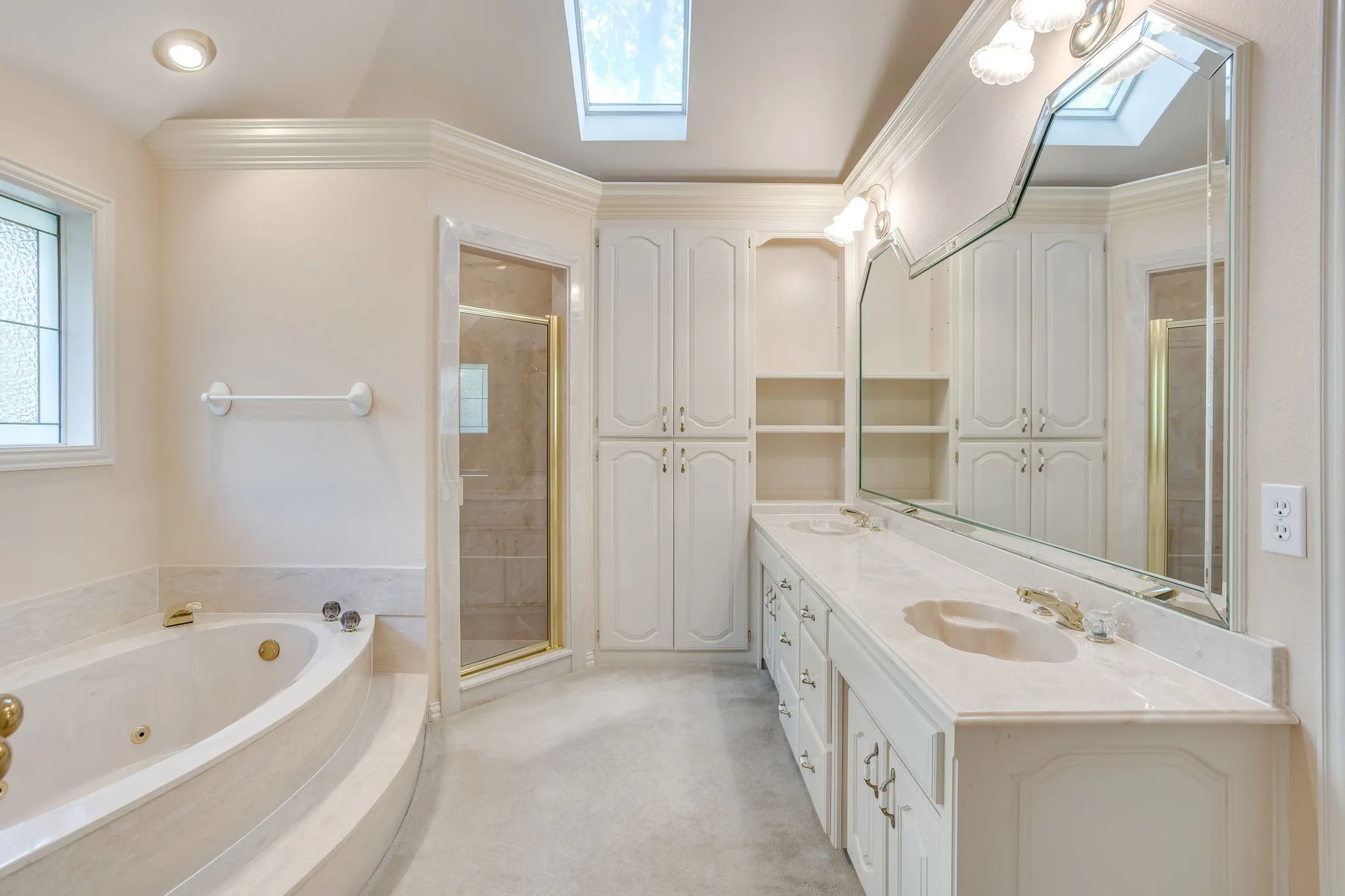 Dual vanity with integrated sinks, built-in cabinetry, jetted soaking tub with steps, glass-enclosed shower, and skylight