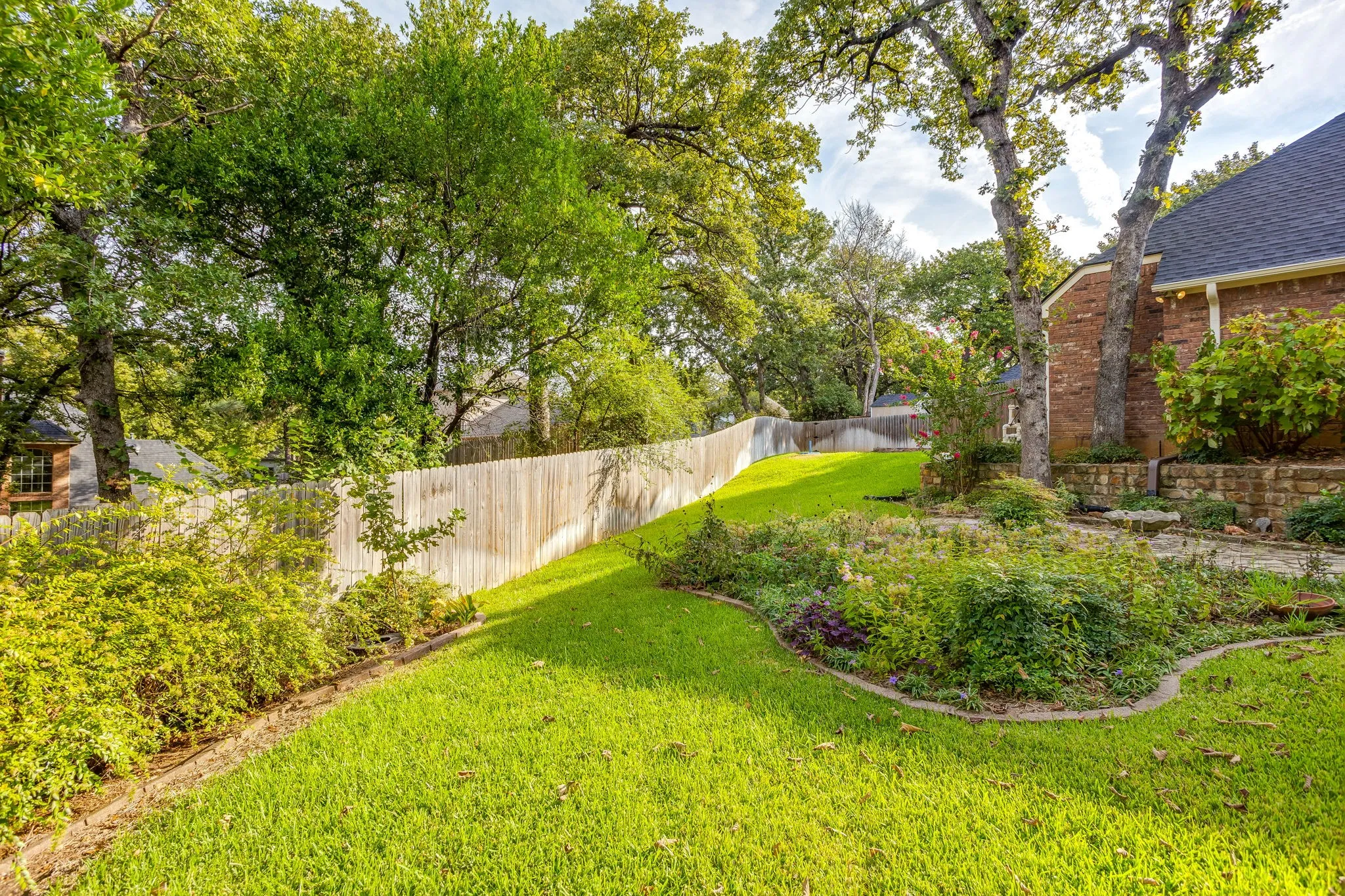 Expansive grassy yard with mature trees, a wooden privacy fence, and manicured landscaping featuring various plantings