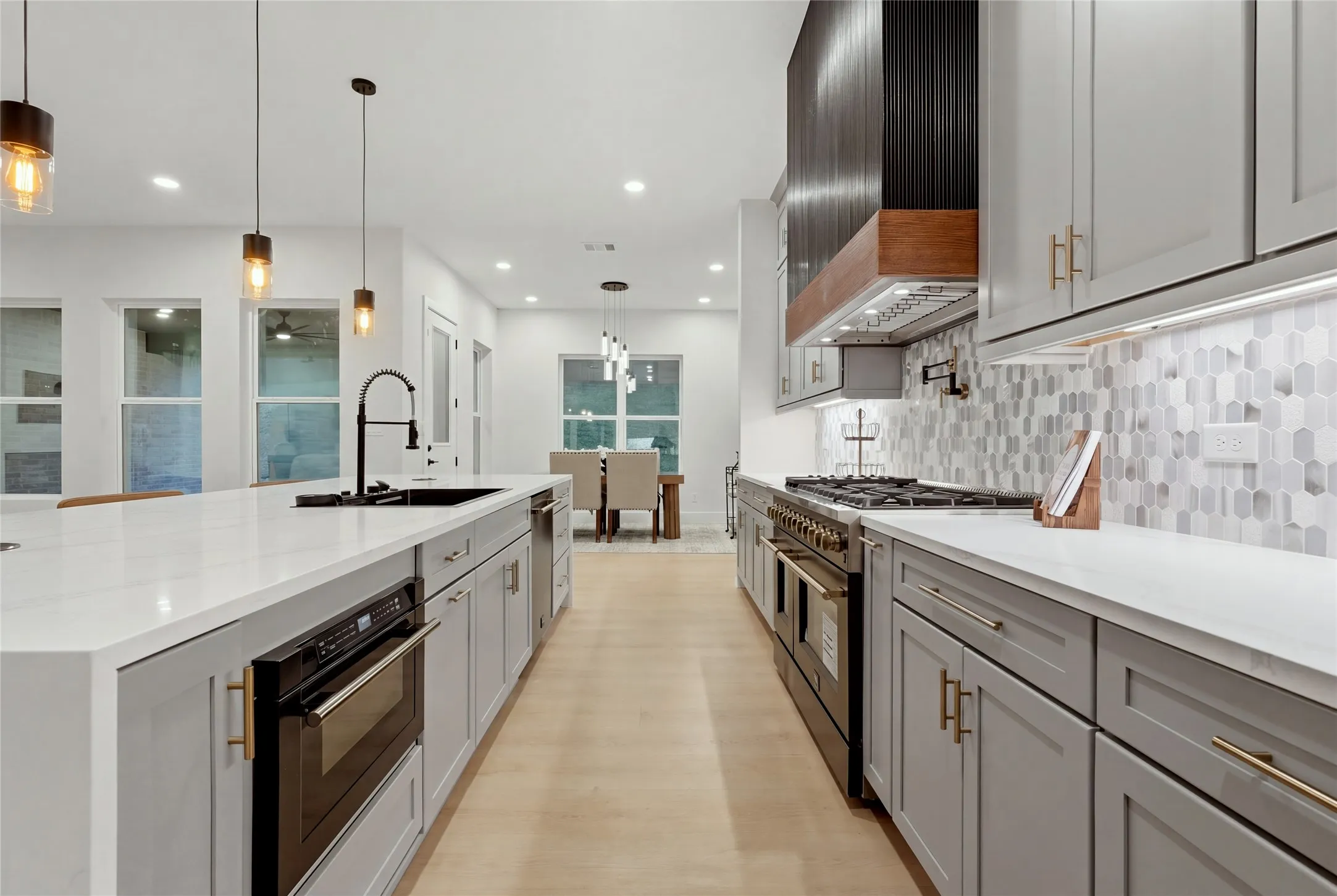 Kitchen featuring light wood-finish flooring, white countertops, gray cabinetry with brass-finish hardware, a large kitchen island with an undermount sink, and stainless steel appliances