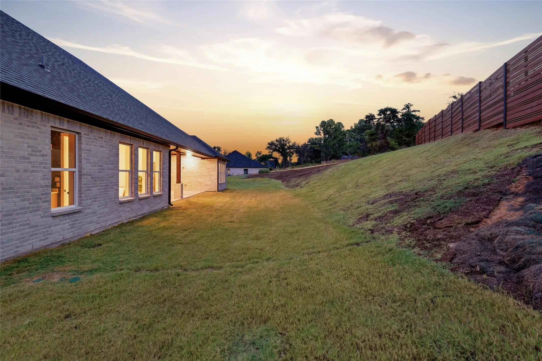 Exterior featuring light-toned brick, multiple windows with white trim, a dark shingle roof, and a wood-slat fence