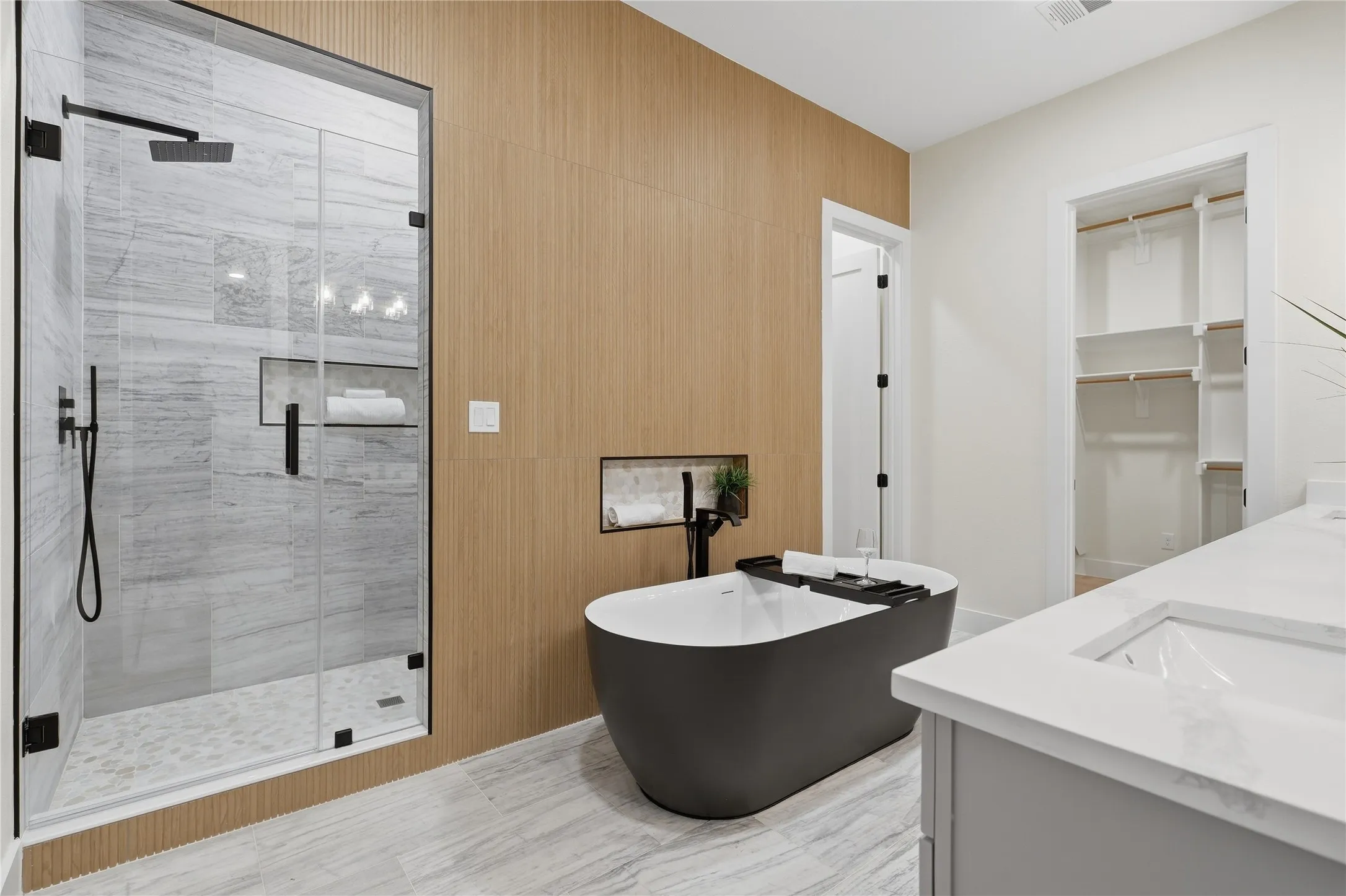 Glass-enclosed shower with a rainfall showerhead and integrated shelving, freestanding soaking tub with a wall-mounted faucet, dual vanity with undermount sinks and white countertops, wood-finish accent wall, and a custom closet system