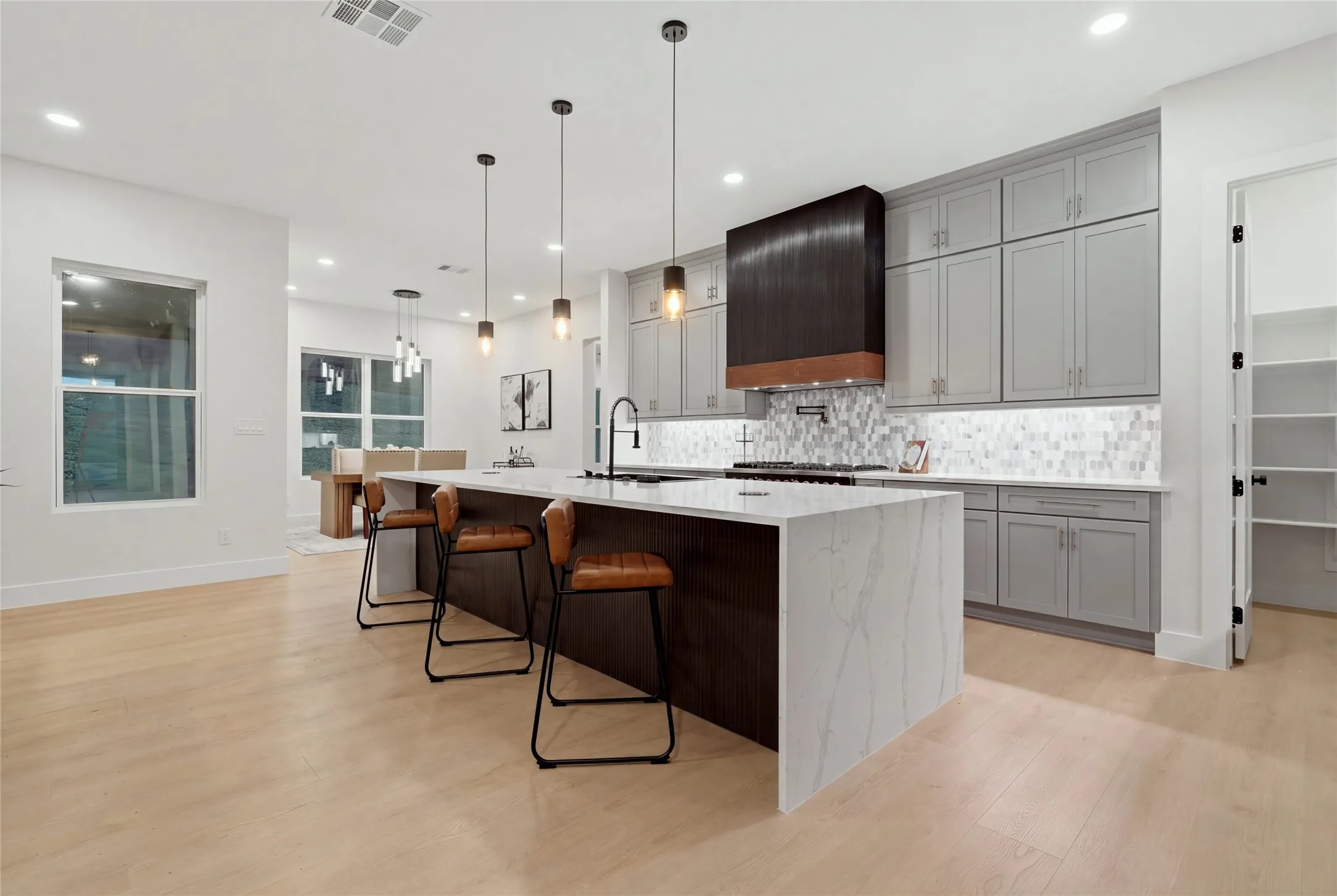 Open-concept kitchen with a large waterfall island, light wood-finish flooring, recessed lighting, and a walk-in pantry
