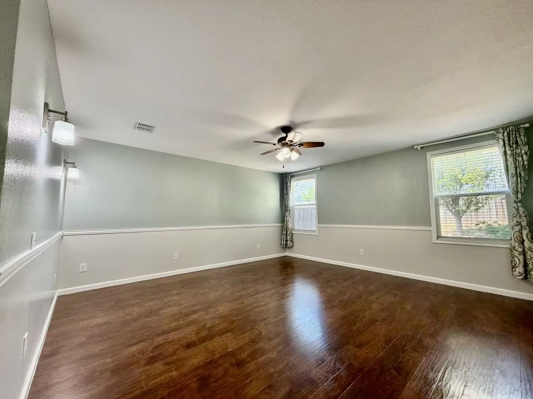 Wood-finish flooring, two large windows, ceiling-mounted fan with integrated lighting, two-tone wall treatment, and wall-mounted sconces