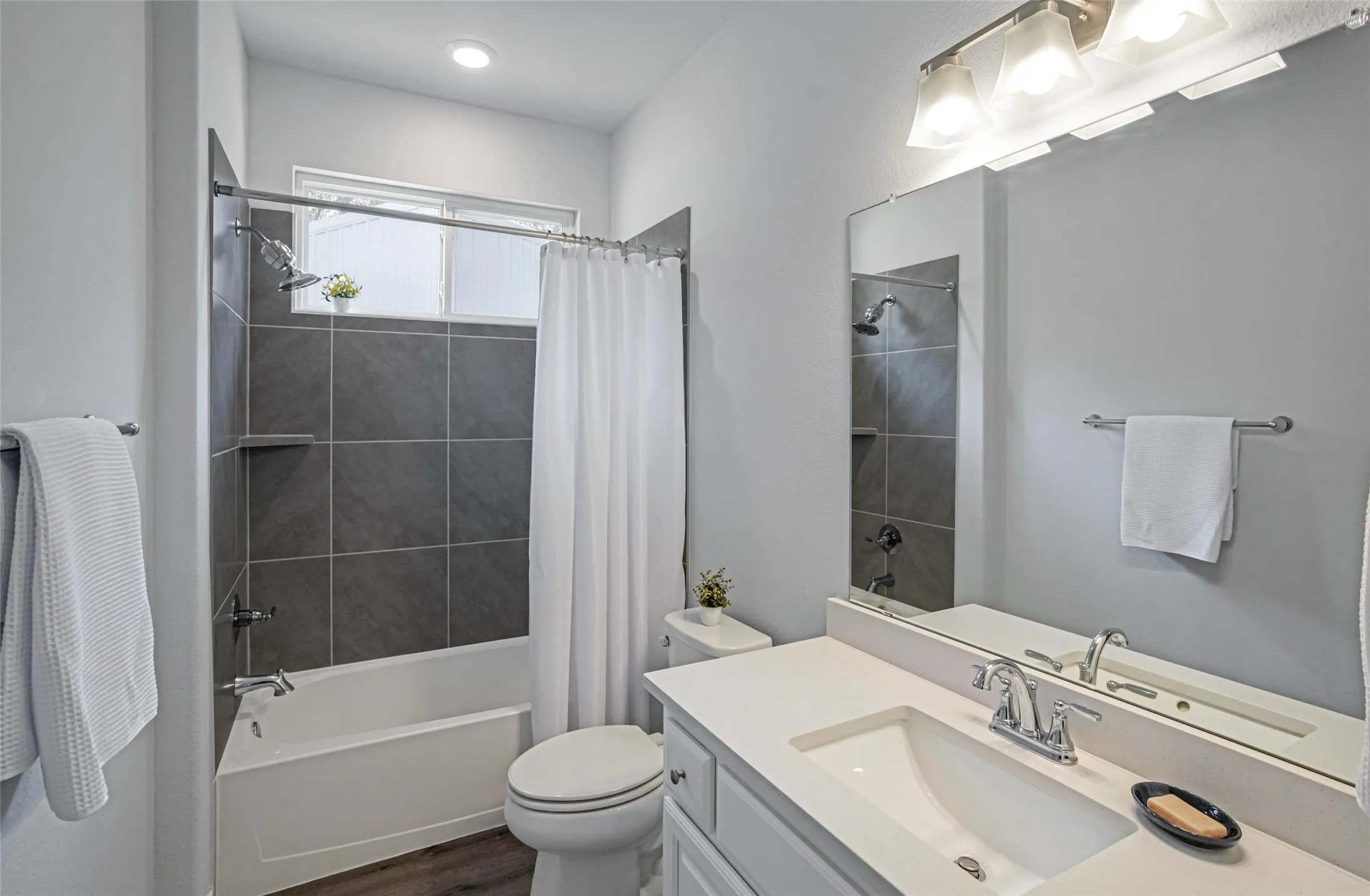 Bathtub with dark gray tile surround, shower with built-in shelf, single vanity with integrated sink, chrome fixtures, and wood-finish flooring