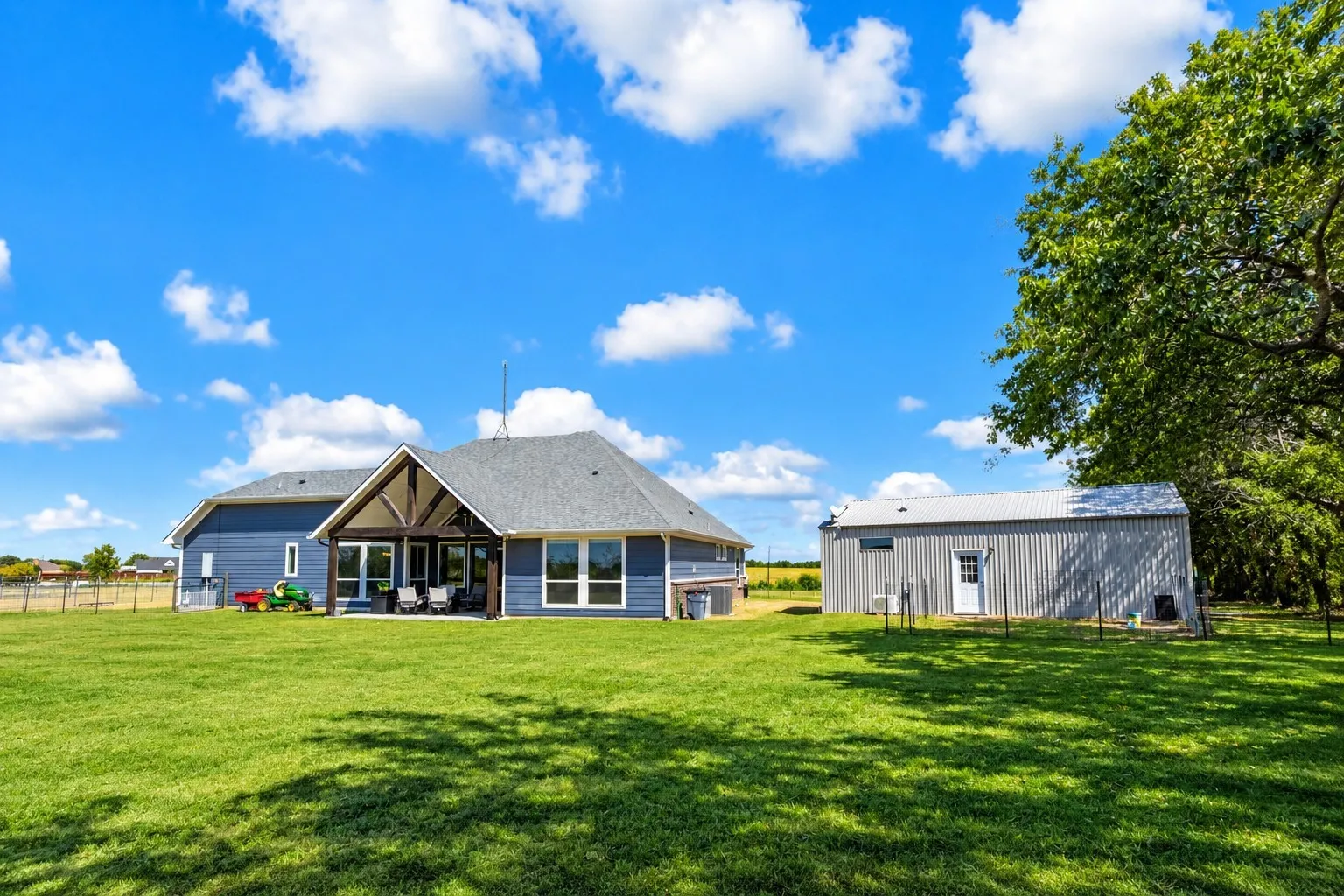 Expansive green lawn with a blue-sided residence, a covered outdoor living area featuring a vaulted wood-beam ceiling, and a detached corrugated metal outbuilding
