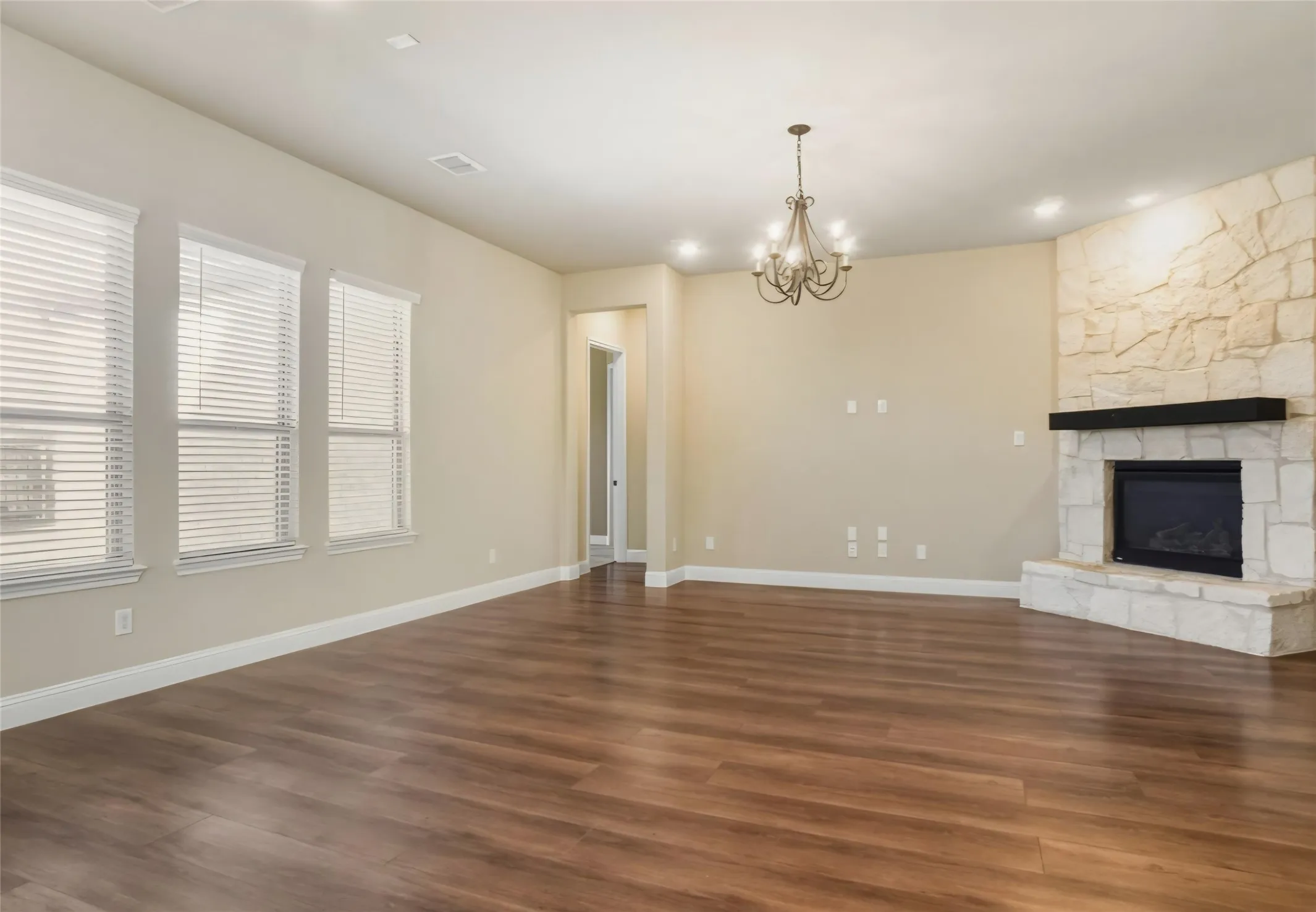Spacious room featuring wood-finish flooring, a stone-clad fireplace with a dark wood mantel, multiple windows with blinds, recessed lighting, and an overhead chandelier