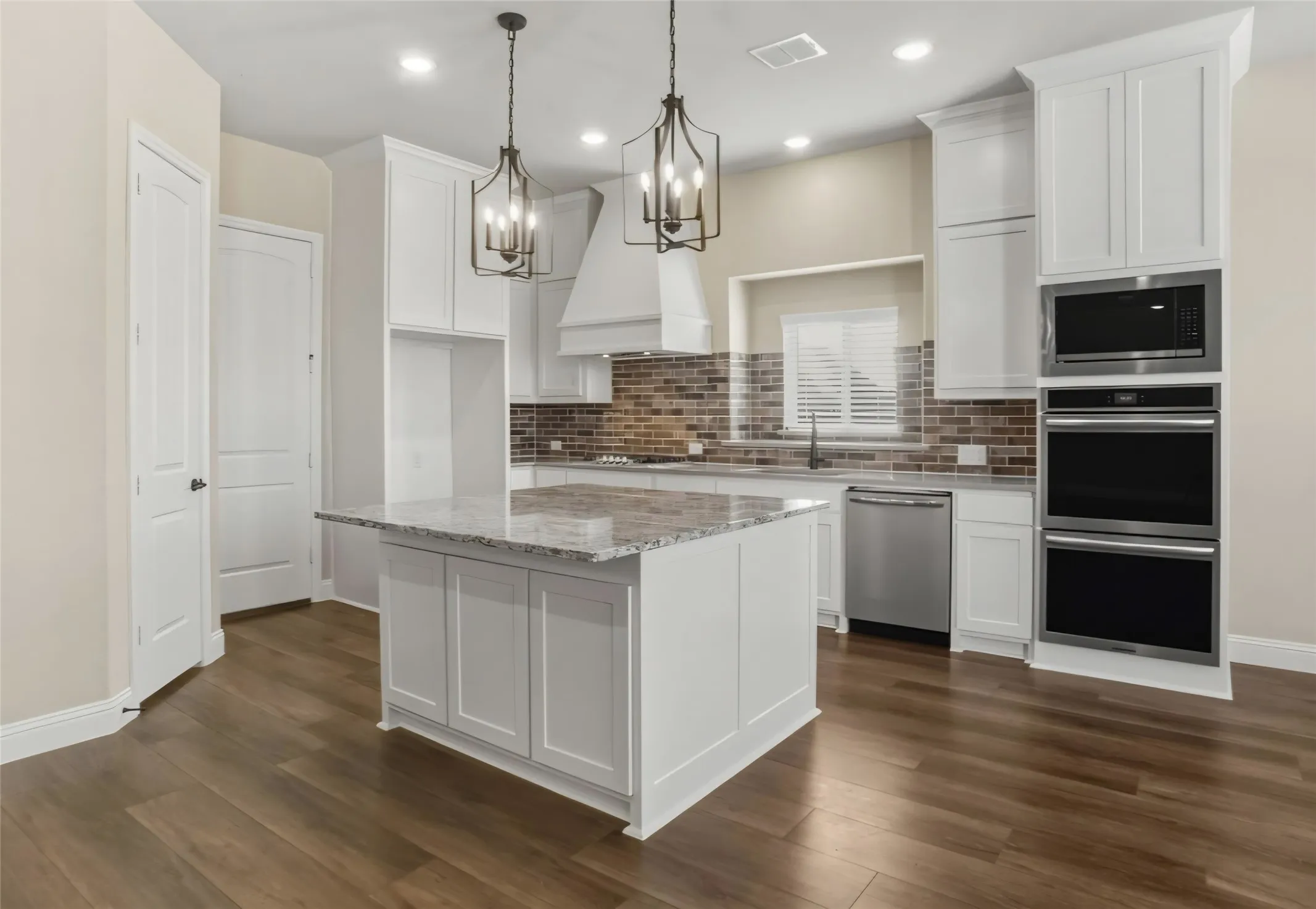 Kitchen with wood-finish flooring, white cabinetry, a central island with a stone countertop, a brick-patterned backsplash, and stainless steel appliances including a built-in microwave, double ovens, and a dishwasher