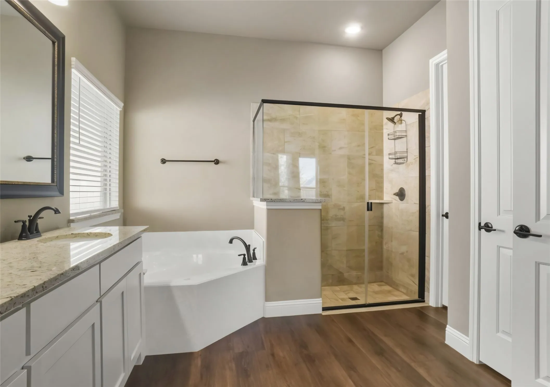 Dual sink vanity with stone countertop, built-in soaking tub, walk-in shower with tile surround, and dark wood-finish flooring