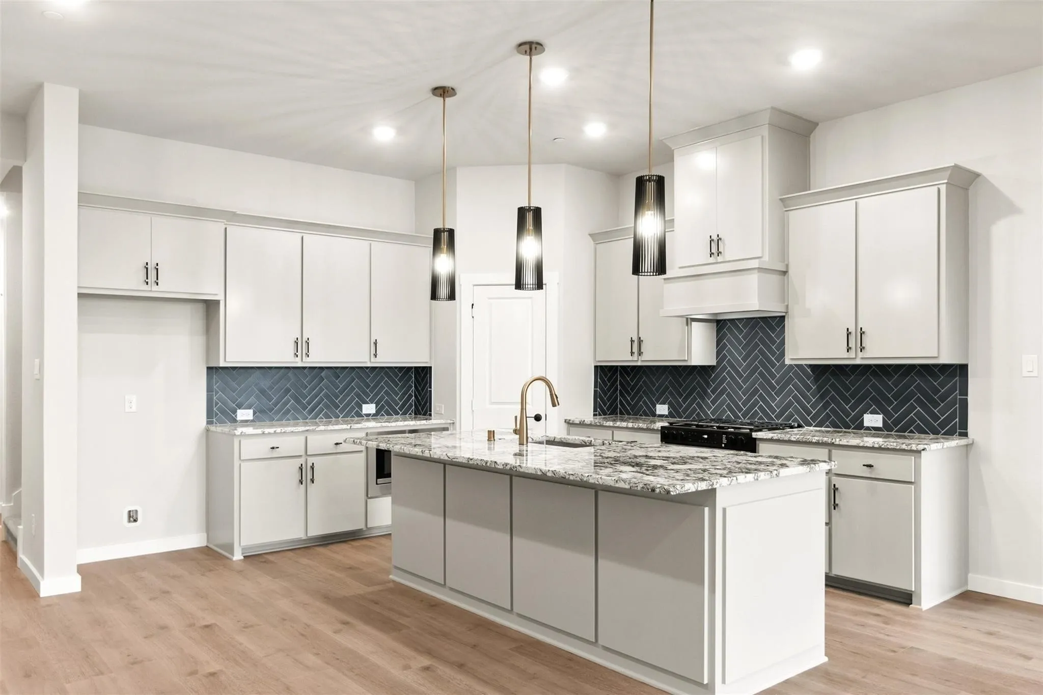 Kitchen with tasteful backsplash, light wood-type flooring, a center island with sink, hanging light fixtures, and light stone counters