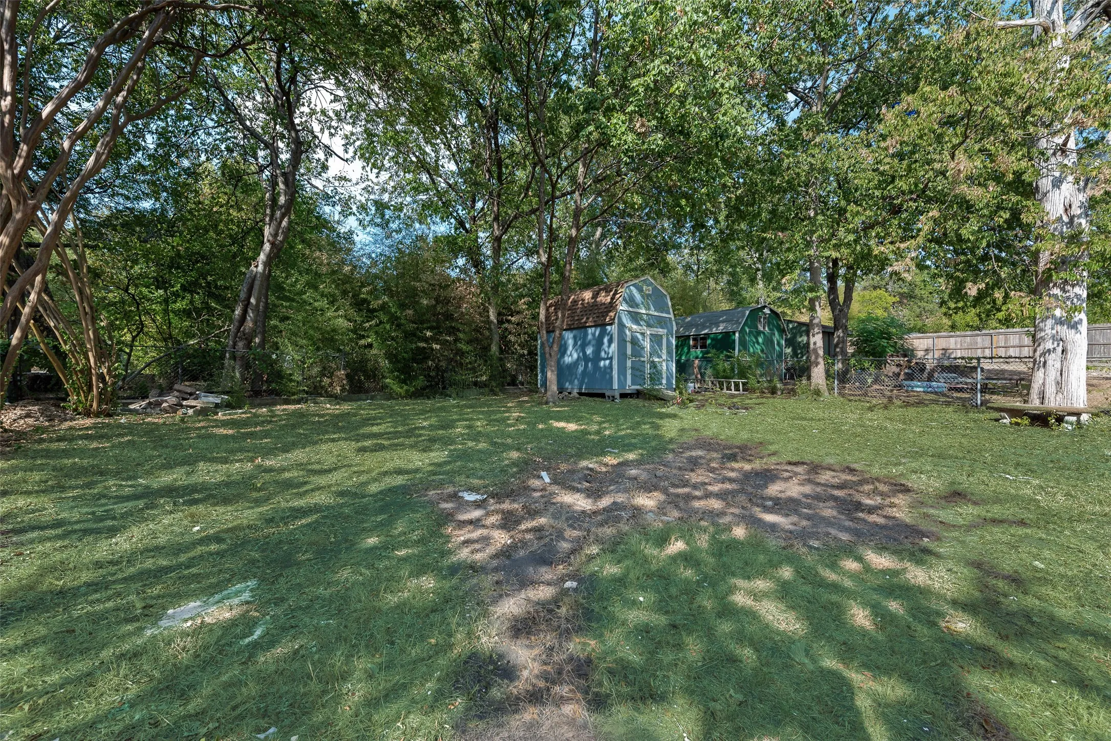 Expansive green lawn with mature trees, featuring a light blue shed with a red roof, a dark green shed, and a wood fence