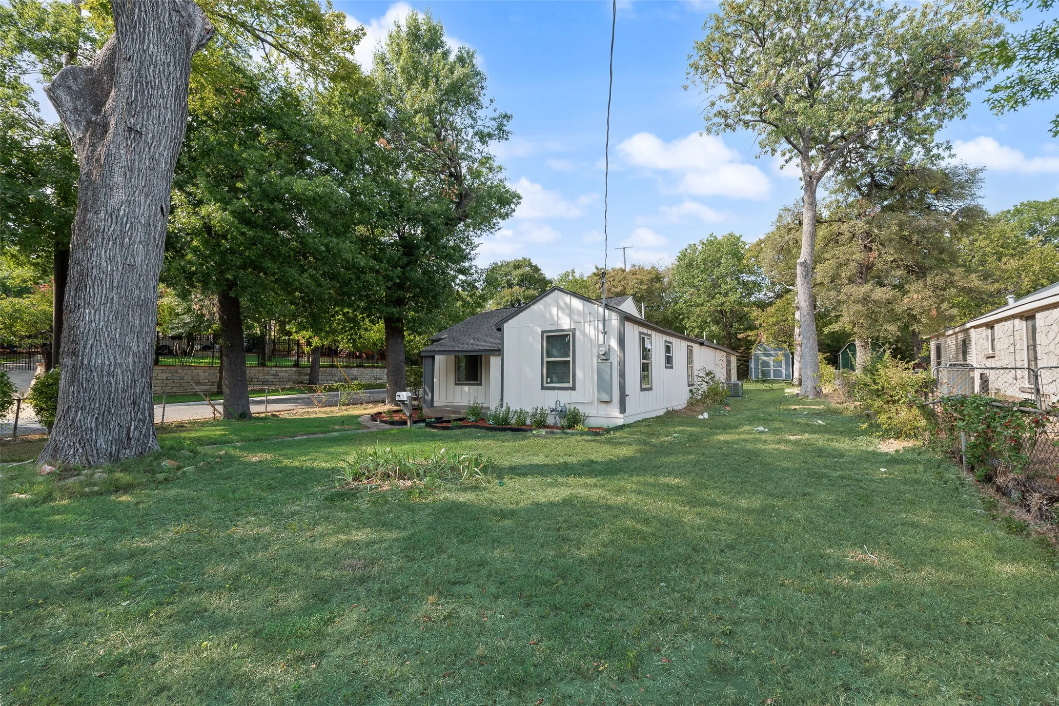 Expansive grassy yard with mature trees, a single-story residence featuring white siding and dark trim windows, and a partial chain-link fence