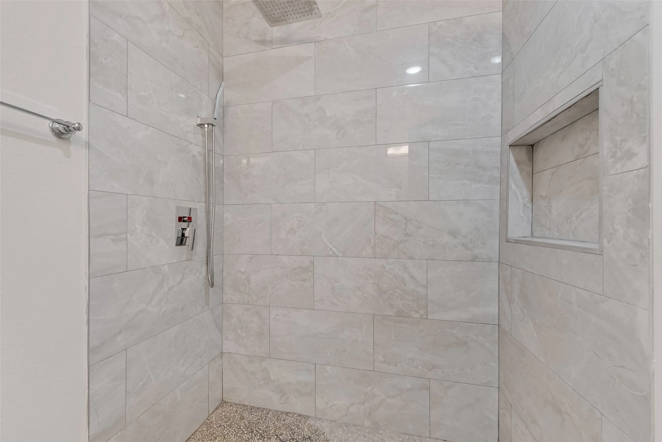 Shower featuring large format wall tiling, a built-in shower niche, a rain-style showerhead, a handheld shower fixture, and a pebble-style floor