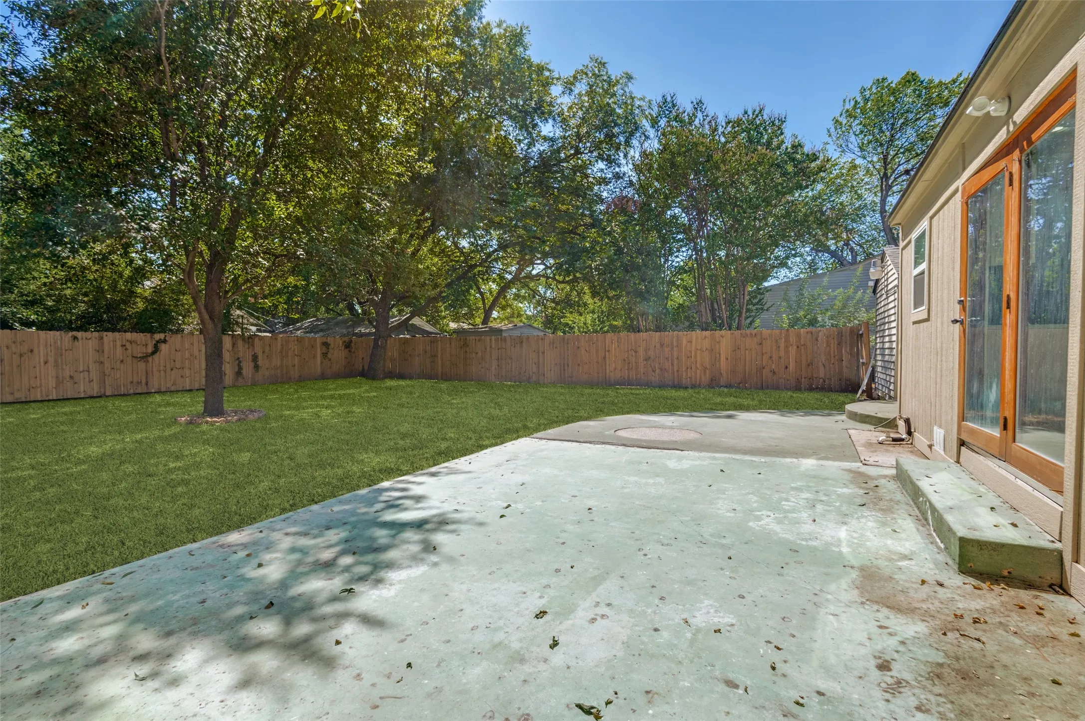 Expansive backyard featuring a manicured lawn, mature trees, wood fence, and a concrete patio
