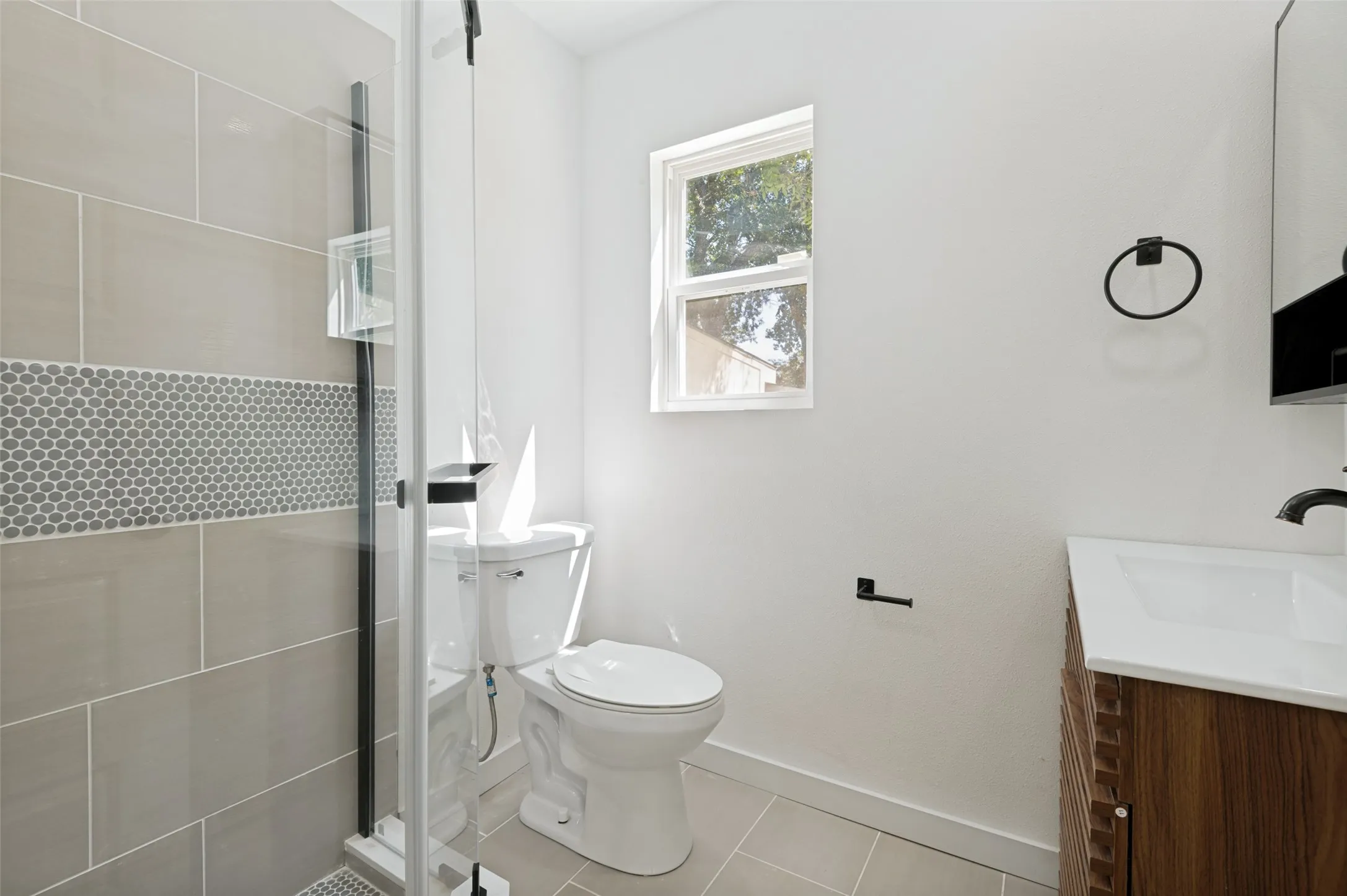 Glass-enclosed shower with gray tiled walls and a mosaic accent band, complemented by a wood-finish vanity with an integrated sink and black fixtures