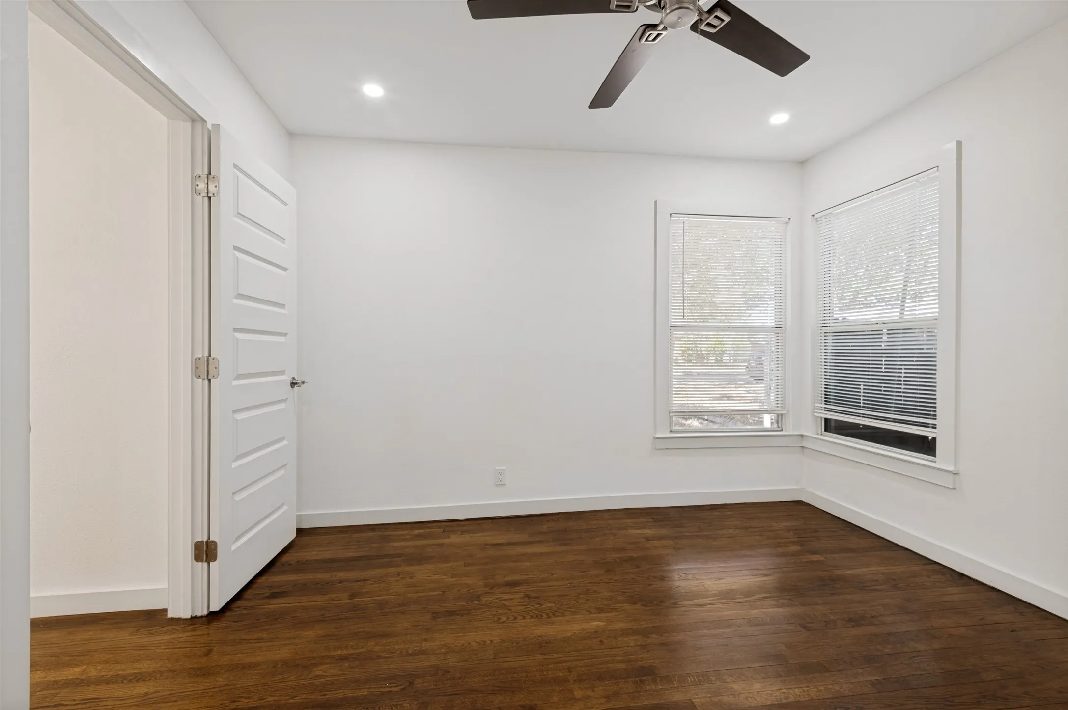 Recessed lighting, wood-finish flooring, two windows with blinds, a ceiling fan, and a paneled door
