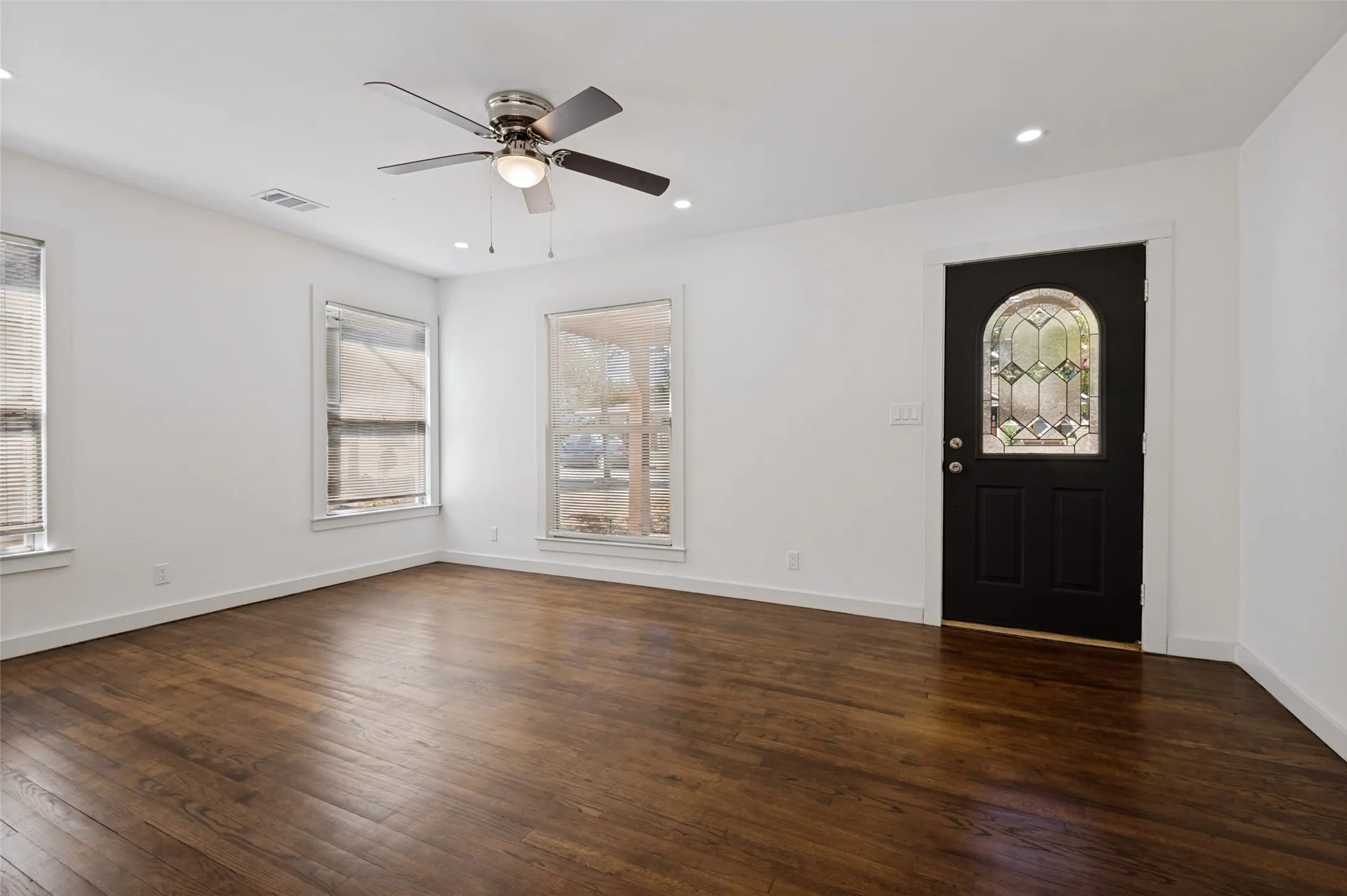 Rich wood-finish flooring, bright white walls, multiple windows, recessed lighting, and a decorative glass entry door