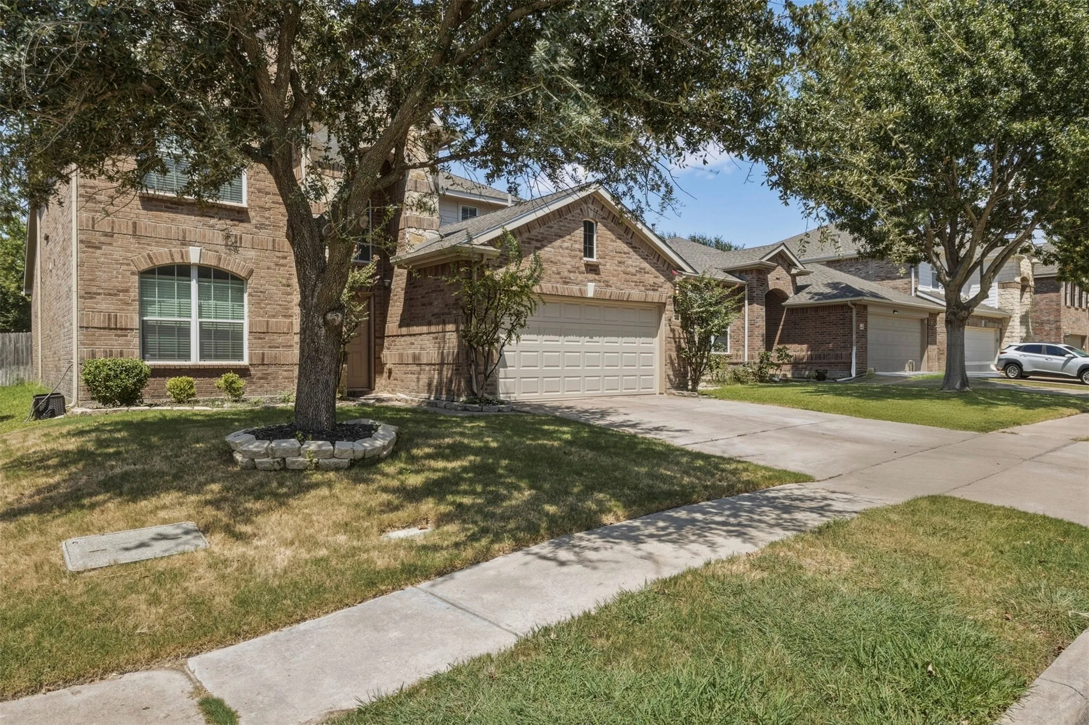 Brick exterior residence featuring an arched window, a two-car garage, and mature trees