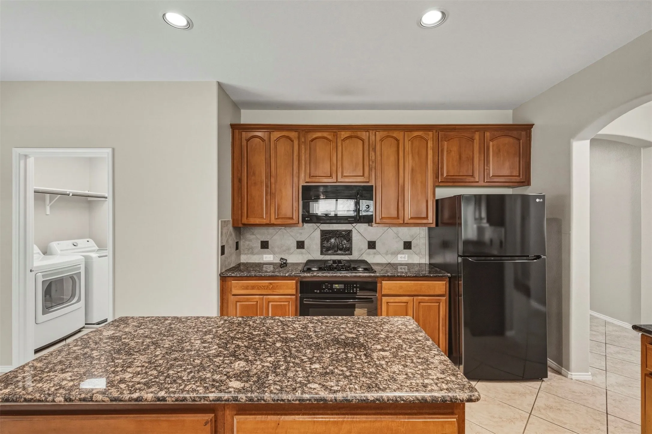 Kitchen featuring recessed lighting, a stone-finish island countertop, wood cabinetry, a tiled backsplash, and black appliances