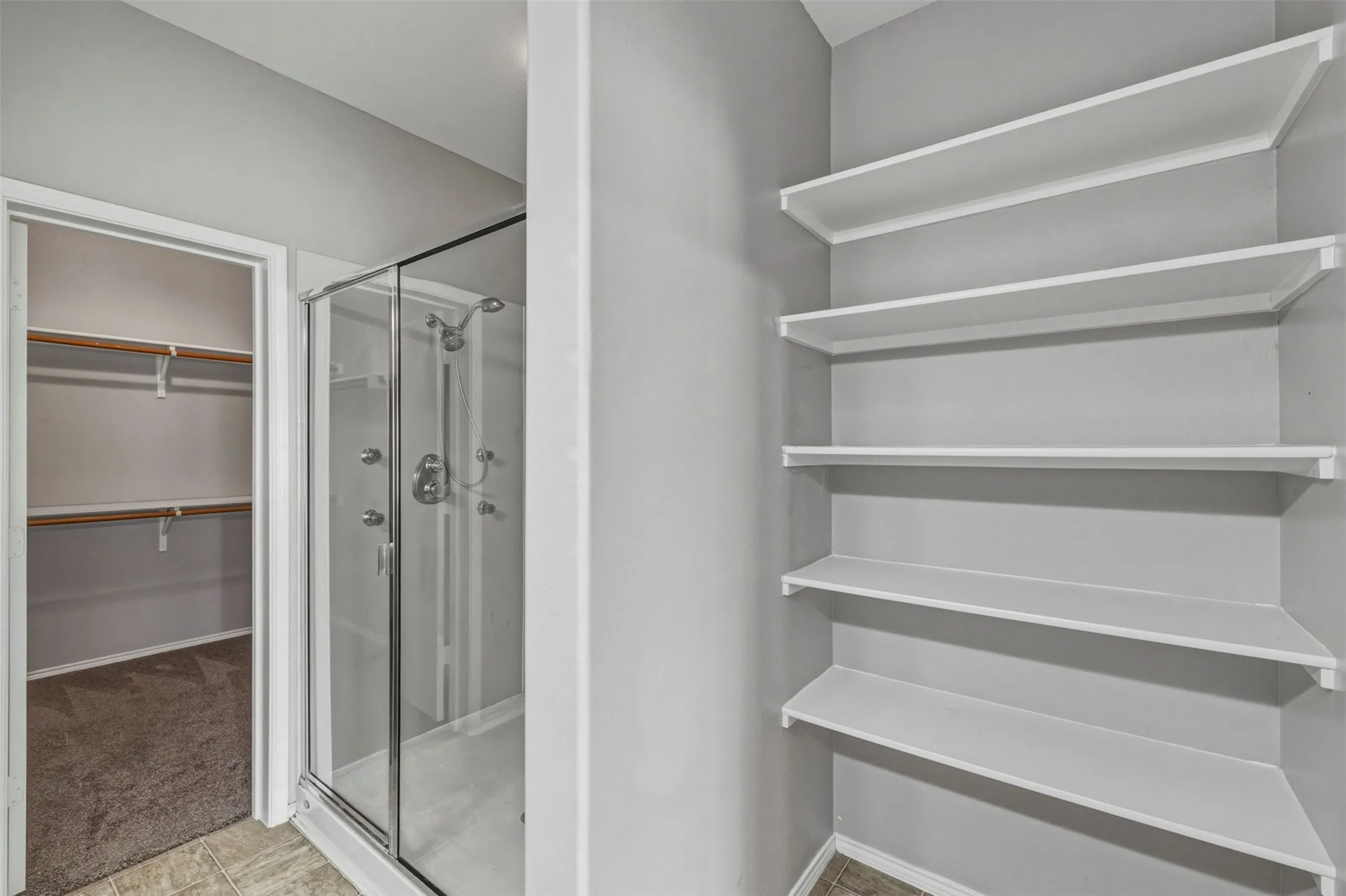 Glass-enclosed shower with multiple sprayers, tiled flooring, built-in shelving, and a separate closet with shelving and hanging rods