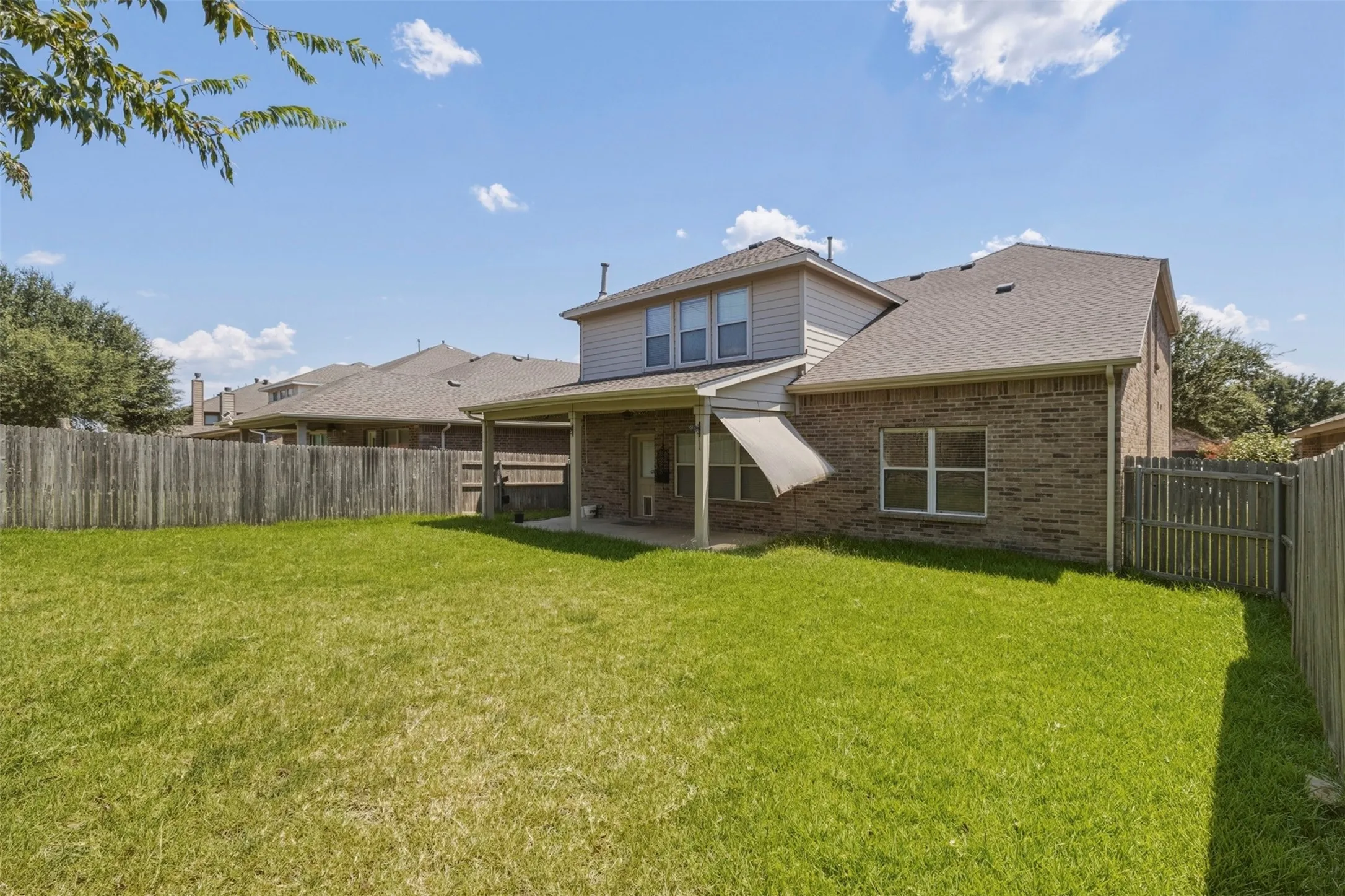 Expansive rear yard featuring green lawn, privacy fencing, a covered patio, and a brick exterior