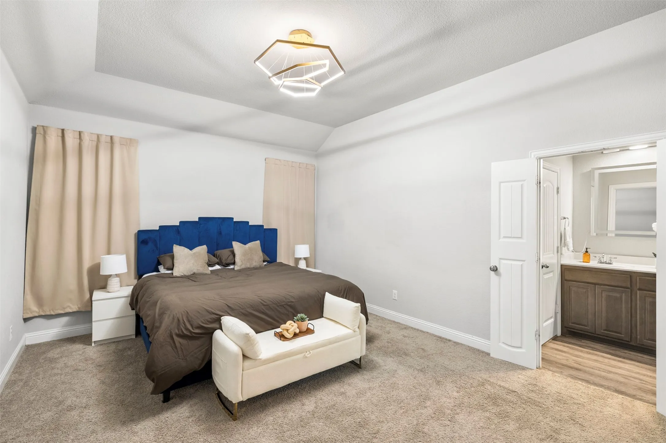 Recessed ceiling with an abstract geometric light fixture, wall-to-wall carpeting, and an ensuite bathroom featuring a vanity with wood-finish cabinetry and a framed mirror