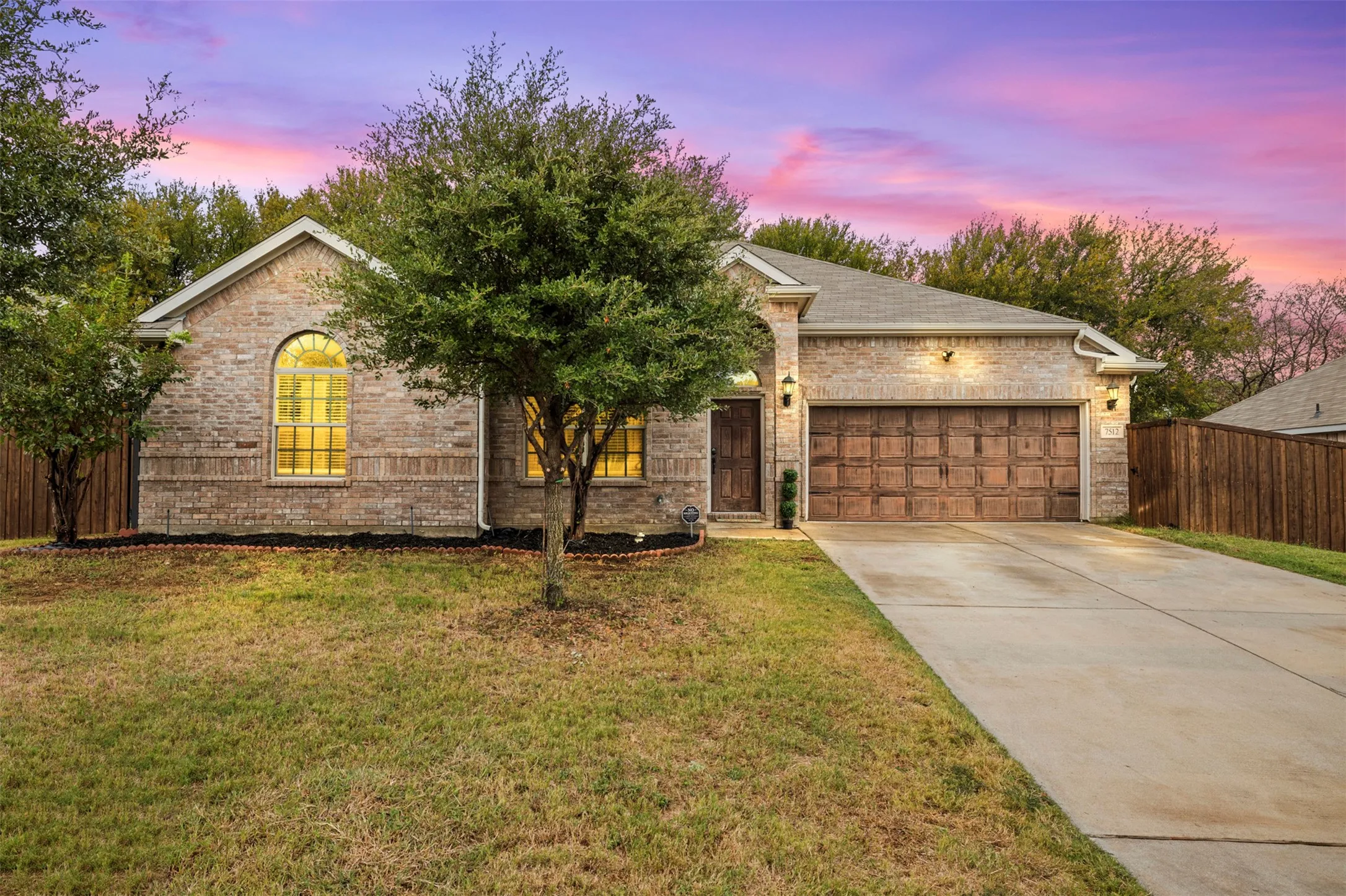 Brick facade, arched window, attached two-car garage with wood-finish door, concrete driveway, and mature landscaping
