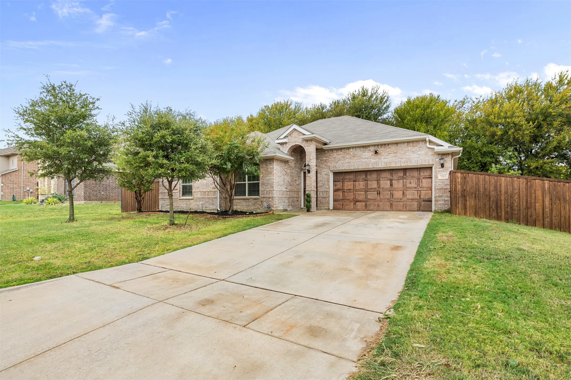 Brick exterior, attached garage with wood-finish door, concrete driveway, and established landscaping