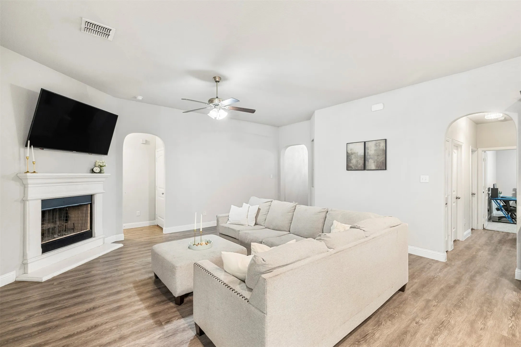 Wood-finish flooring throughout, featuring a ceiling fan with integrated lighting, an arched doorway, and a corner fireplace with a white mantel