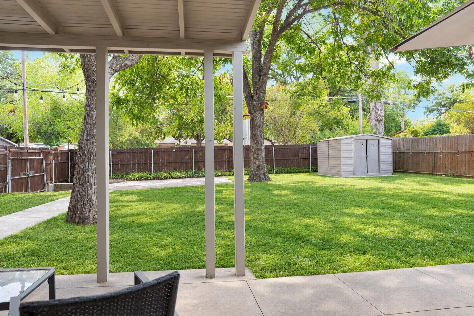 Covered patio with concrete flooring, leading to a lawn, mature trees, and a utility shed