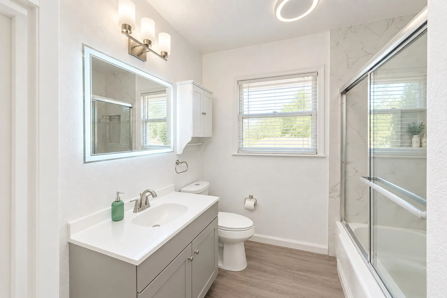 Single vanity with an integrated sink and brushed nickel faucet, a backlit mirror, a built-in wall cabinet, and a shower-over-tub with glass enclosure