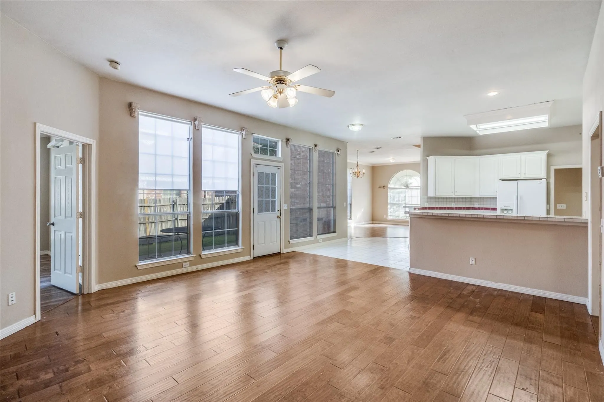 Wood-finish flooring, ceiling fan with light fixture, multiple large windows with blinds, white kitchen cabinetry, and a kitchen island with a tiled backsplash