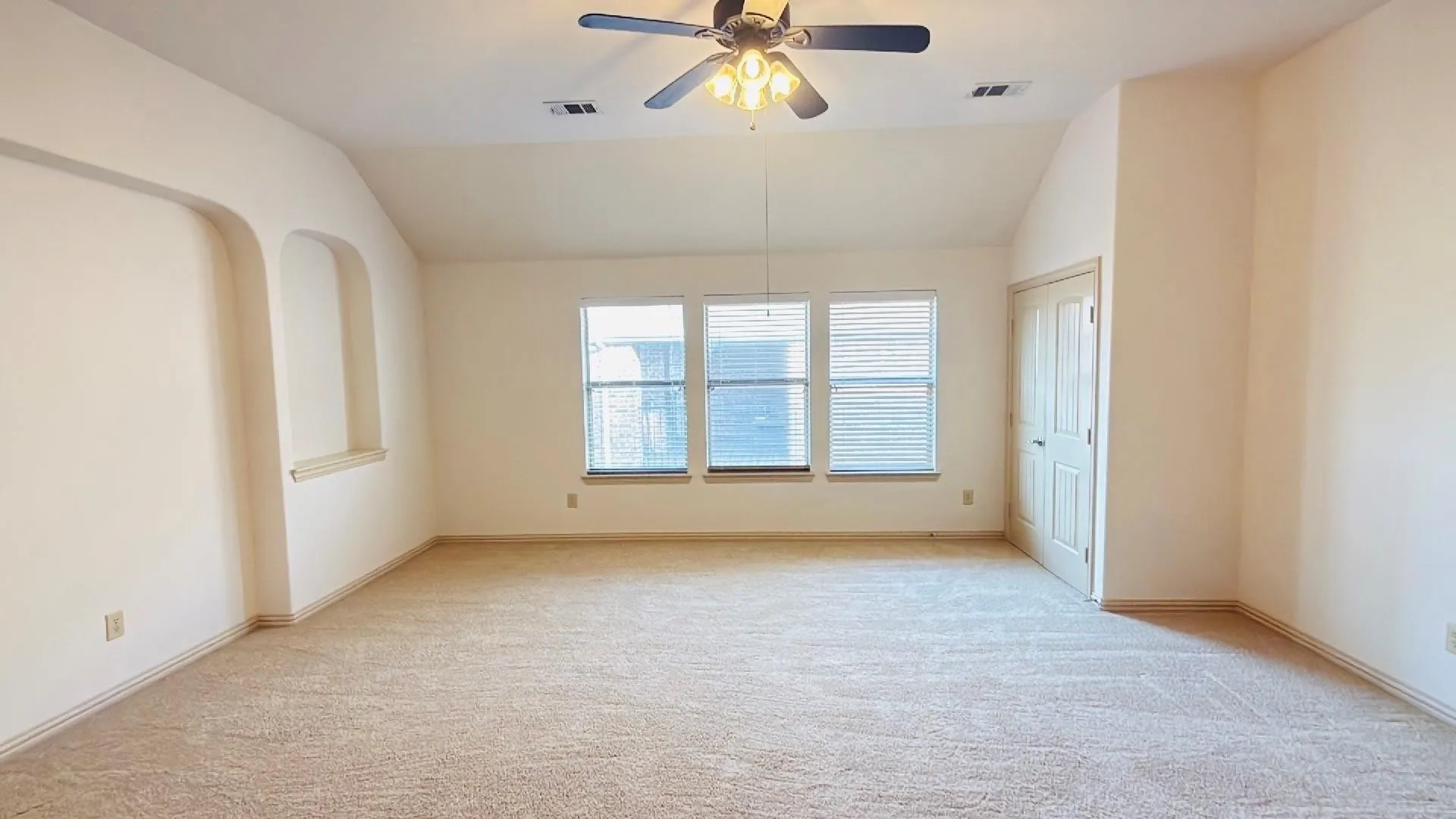 Carpeted room with vaulted ceiling, arched wall alcoves, multiple windows with blinds, and a paneled door