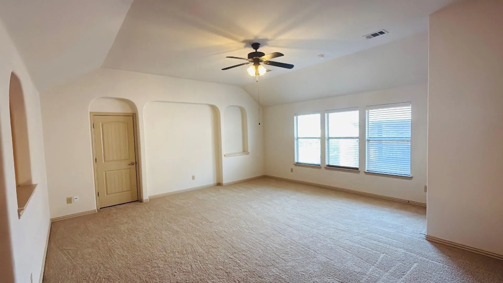 Expansive room featuring vaulted ceilings, a ceiling fan with integrated lighting, three large windows with blinds, built-in wall niches, and neutral carpeting