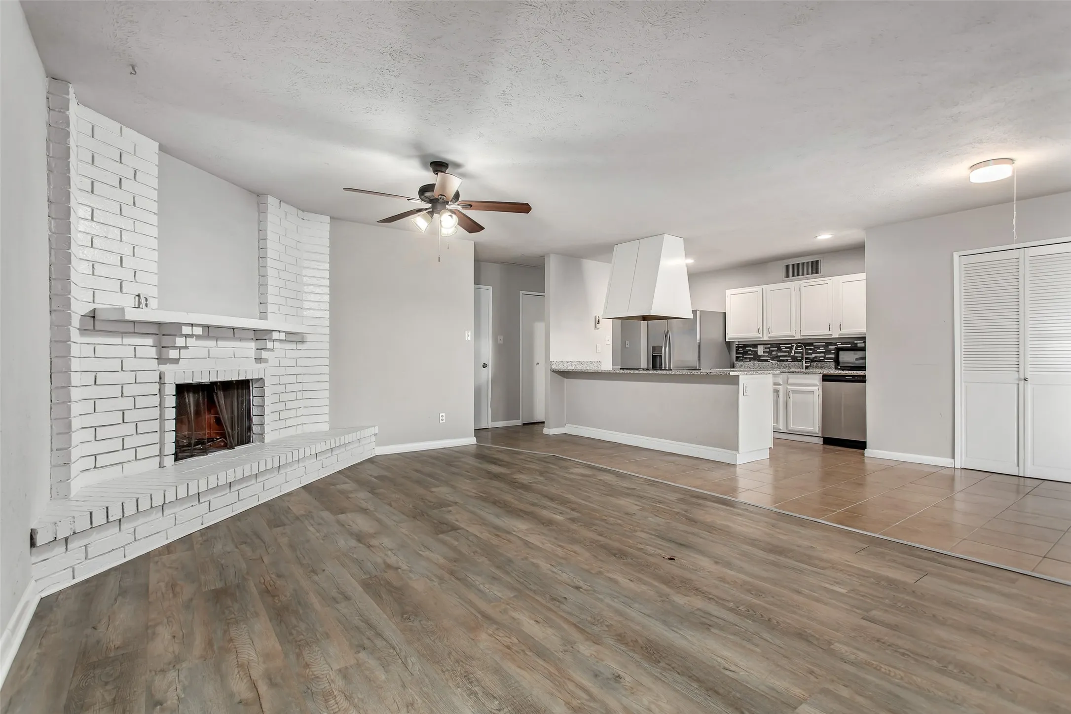Wood-finish flooring, white brick fireplace with mantel, ceiling fan, kitchen with white cabinetry, and stainless steel appliances