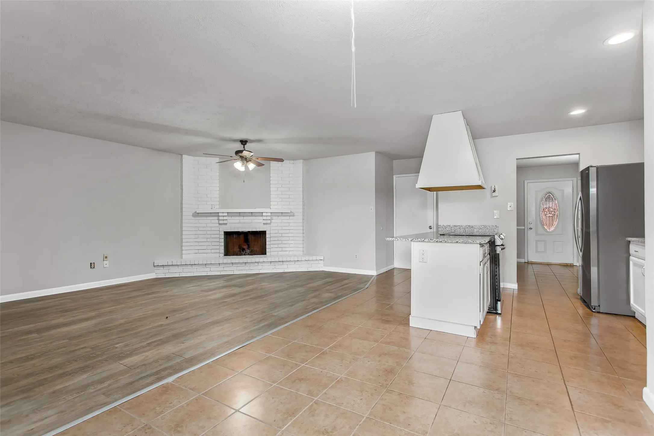 White painted brick fireplace, wood-finish flooring, ceiling fan, granite-finish countertop on kitchen island, and ceramic tile flooring