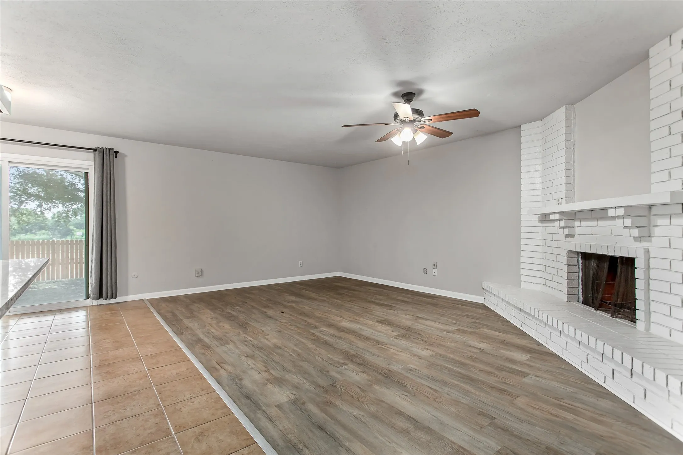 Spacious living area featuring wood-finish flooring, a white brick fireplace with mantel, and a ceiling fan with integrated lighting