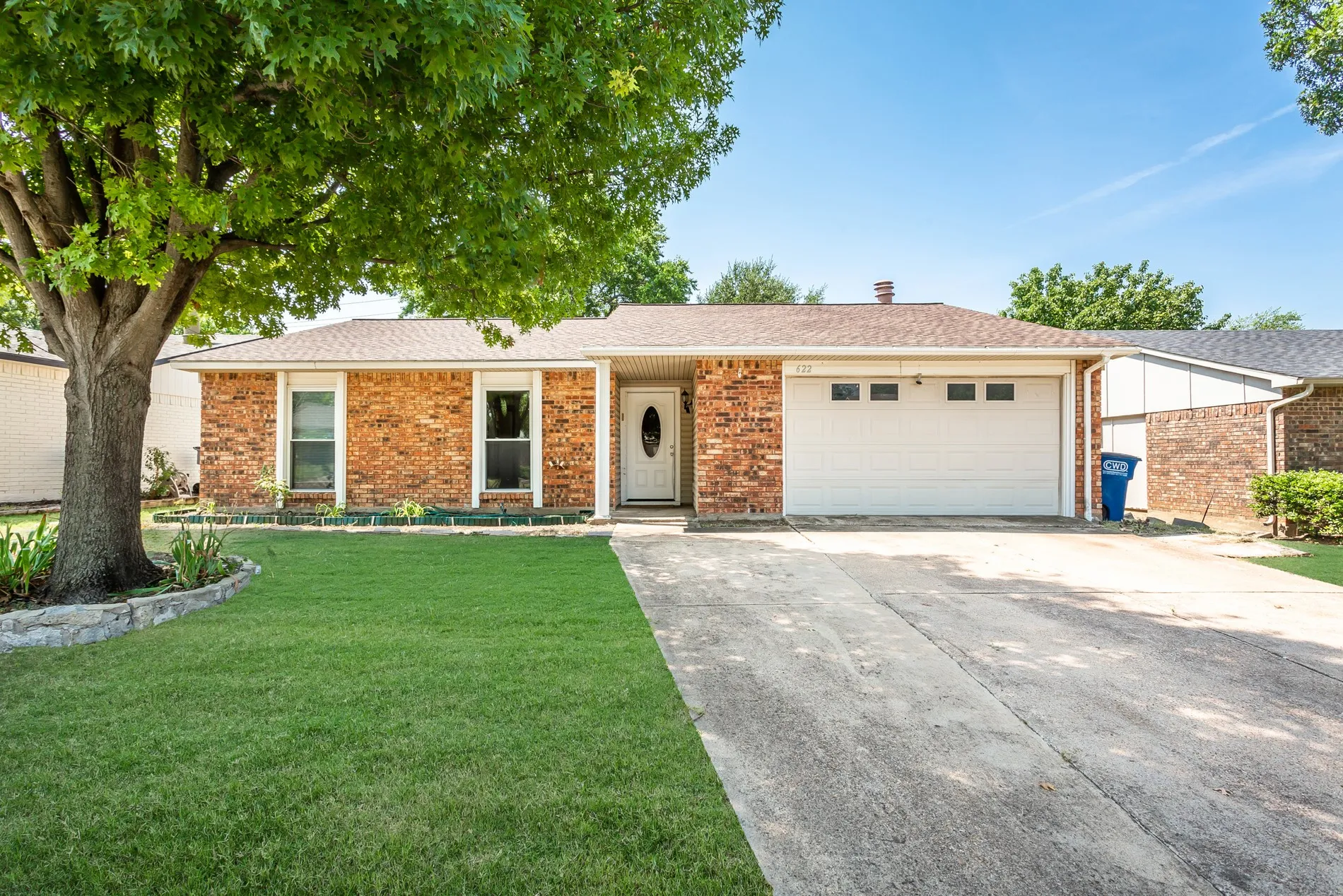 Brick exterior with a white entry door, an attached two-car garage, mature landscaping, and a concrete driveway