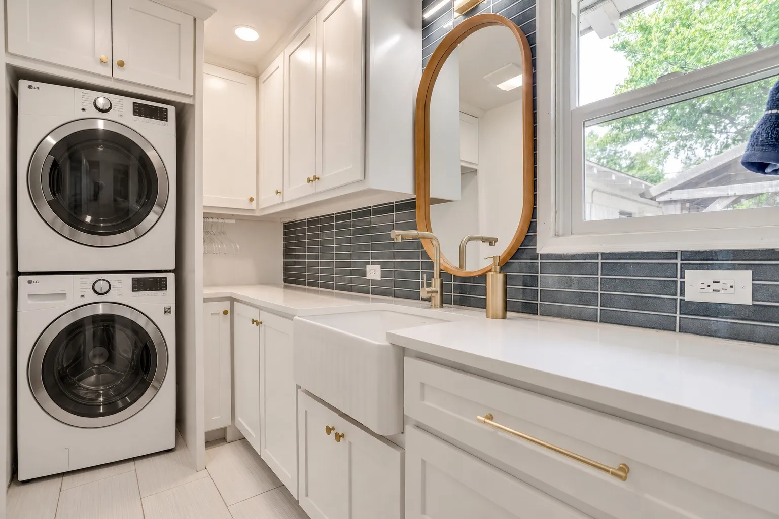 White shaker cabinetry with gold-finish hardware, virtually-staged stacked washer and dryer, farmhouse sink, white countertops, and blue rectangular tile backsplash