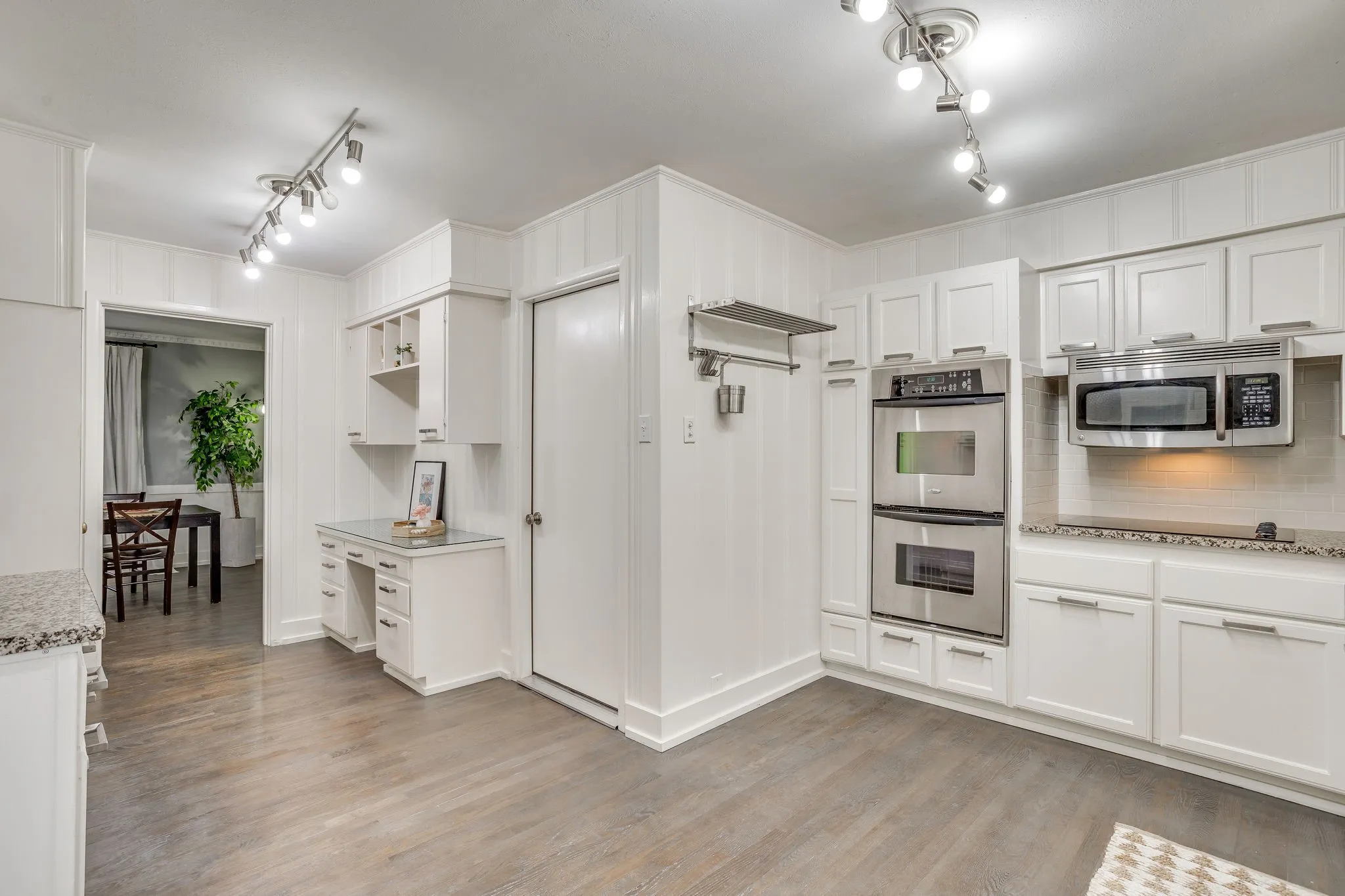 White cabinetry, wood-finish flooring, stainless steel double oven, stainless steel microwave, and track lighting