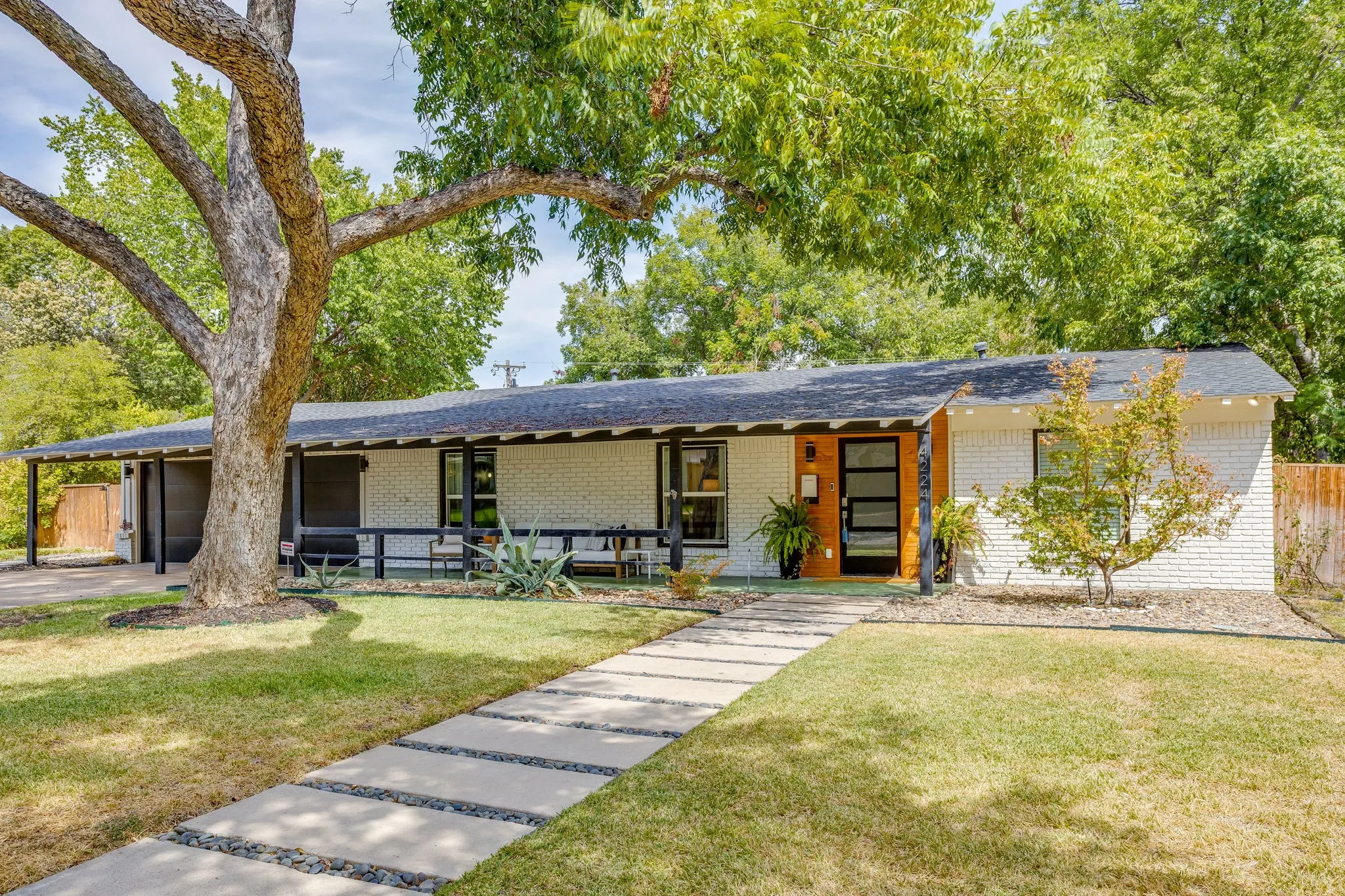 White brick exterior, dark-trimmed windows, covered front porch, paved walkway, mature front yard tree
